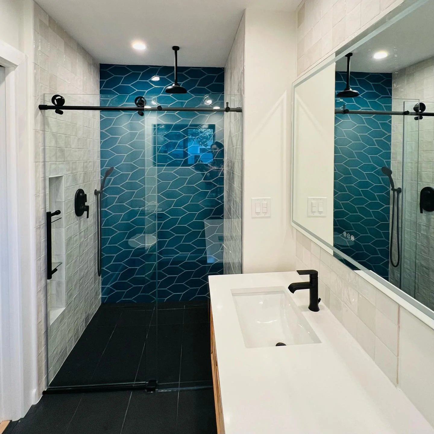 Modern bathroom featuring a walk-in shower with blue hexagon tiles and a black sliding door, a white vanity with a black faucet, and a large mirror. Black fixtures and accents are present throughout.