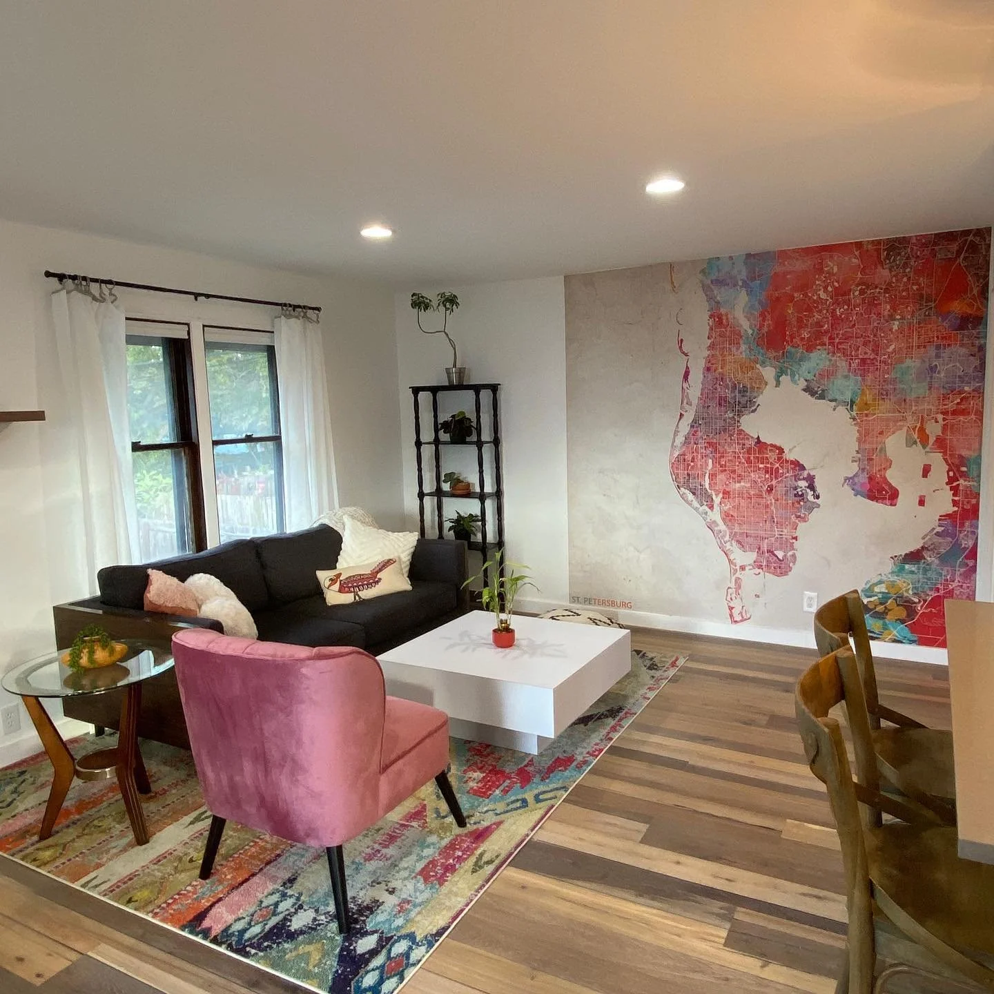 Living room with black sofa, pink armchair, white coffee table, colorful rug, wall mural of a colorful map, window with white curtains, and hardwood floors.