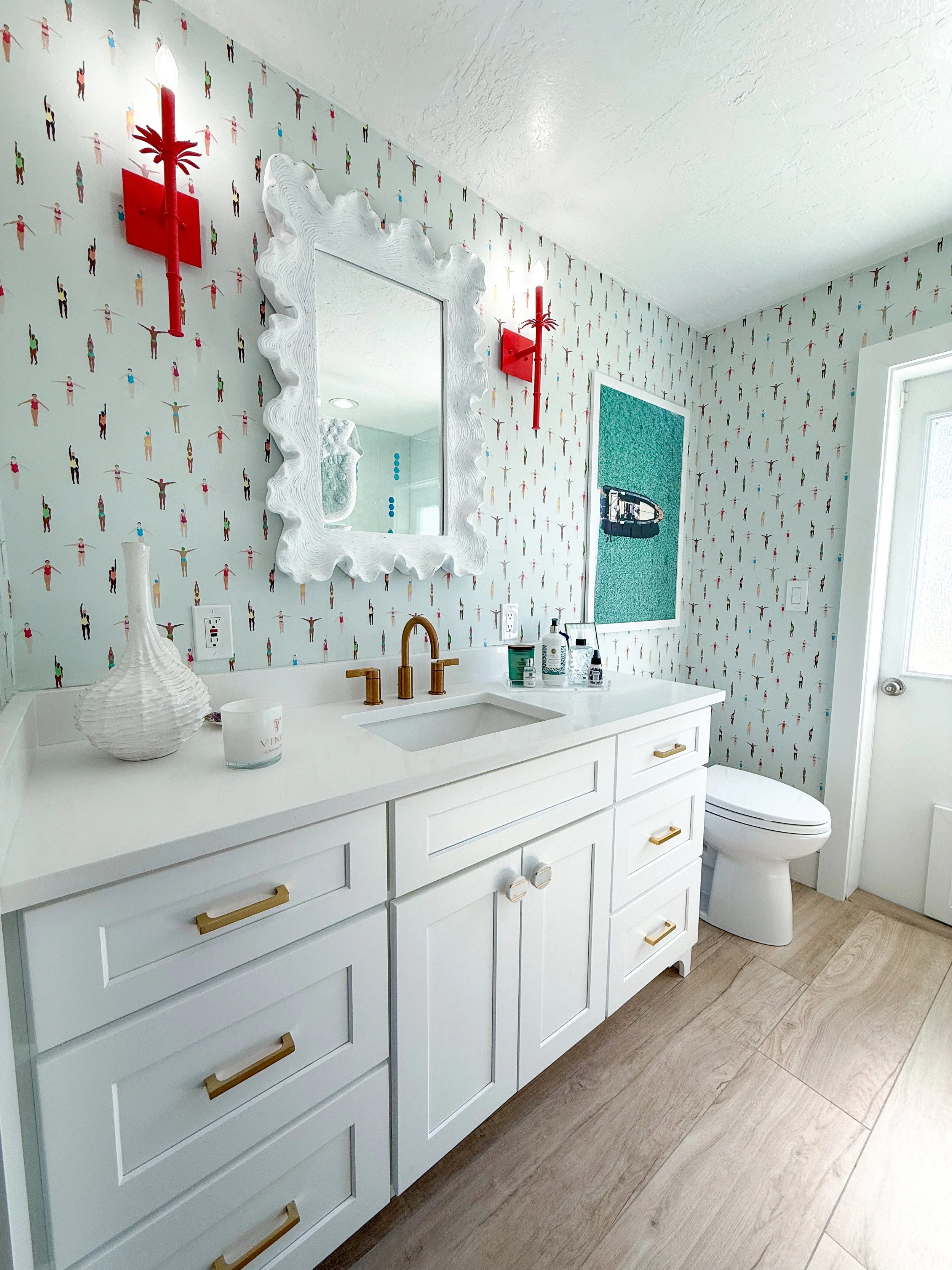 A modern bathroom featuring a white vanity with gold handles, a gold faucet, a mirror with a white decorative frame, red wall sconces, a white vase, and a candle. The walls are decorated with colorful, small, human-shaped figures, and there is a wind