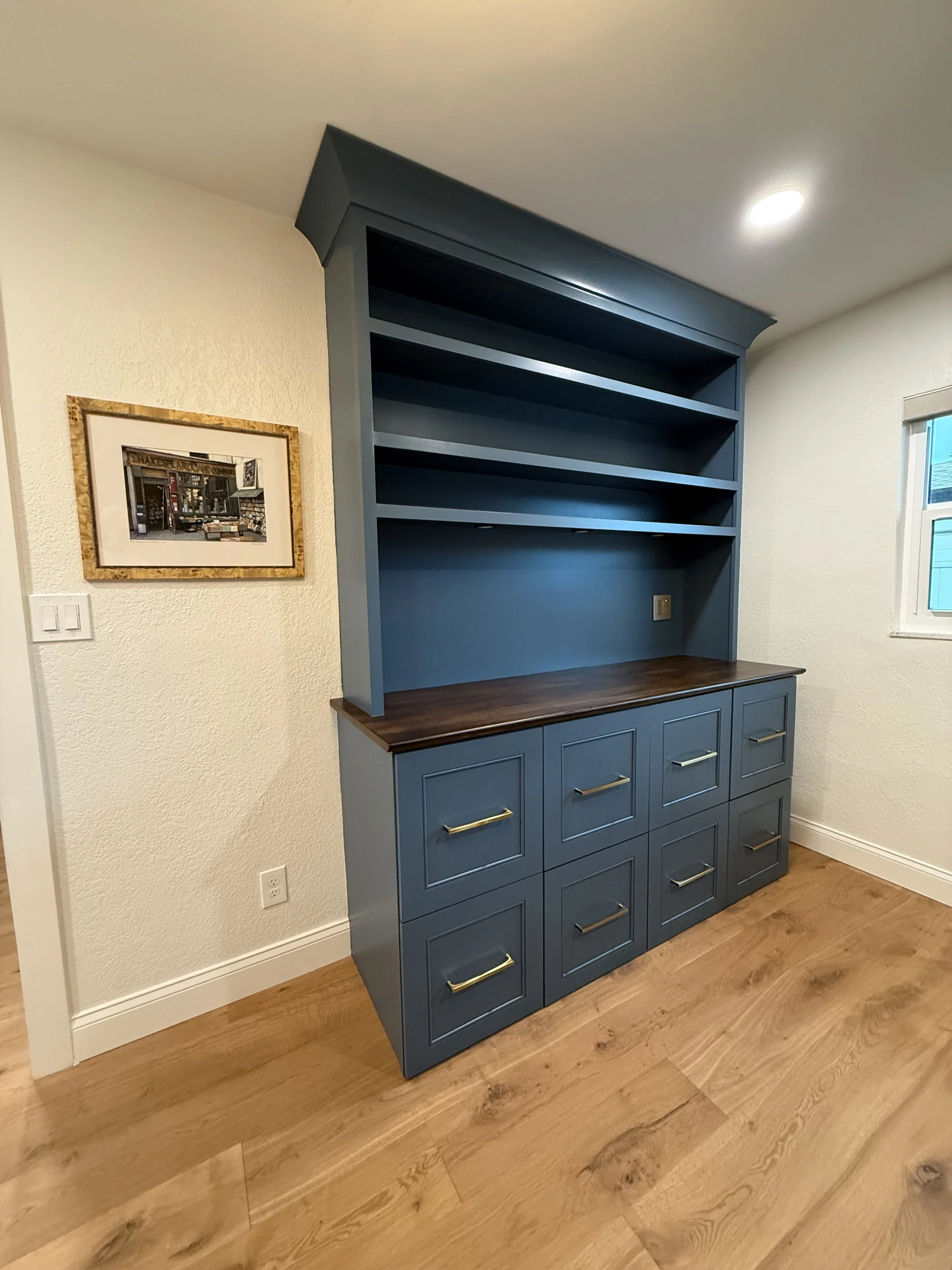 A blue built-in cabinet with open shelves and drawers, featuring a dark wood countertop, situated in a room with white textured walls and hardwood flooring.