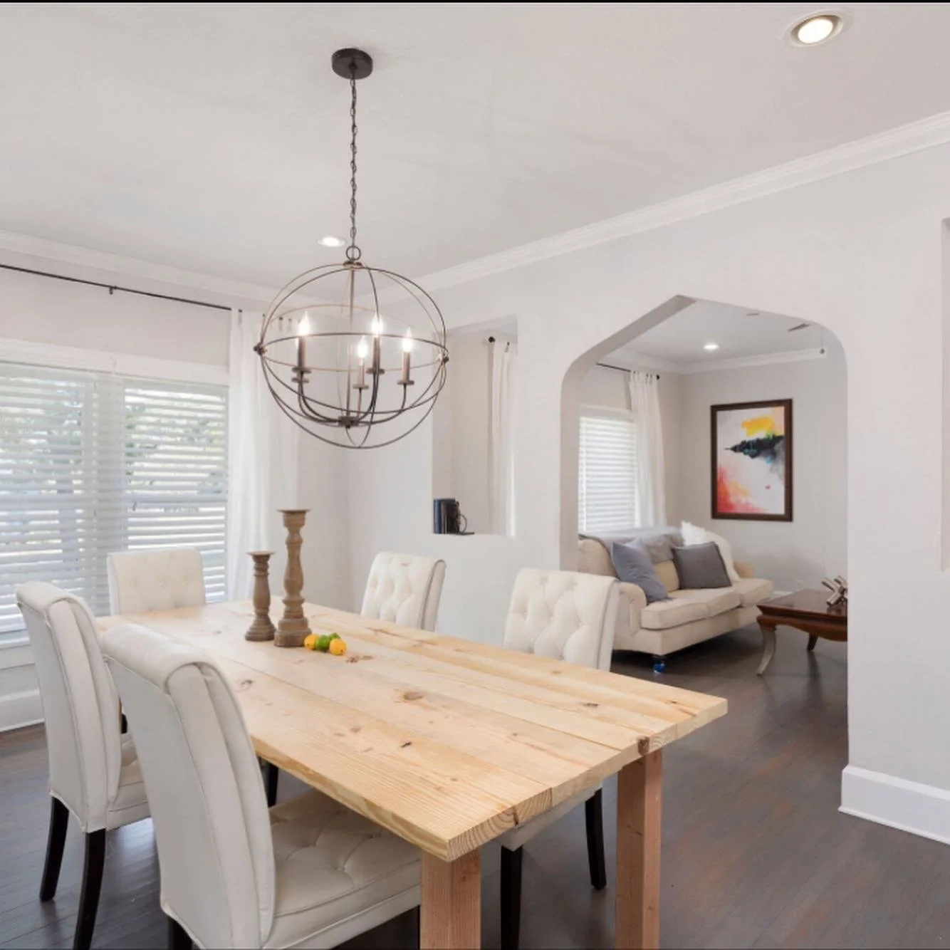 Interior of a dining and living room area with white walls, wooden dining table, beige chairs, chandelier, and a couch with artwork on the wall.