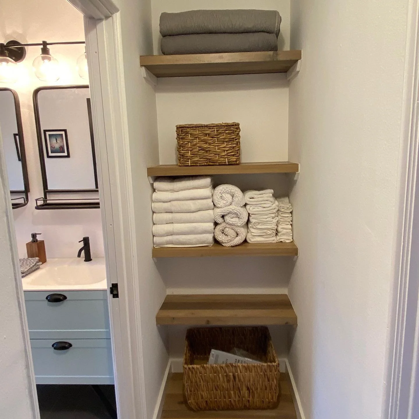 Bathroom linen closet with three wooden shelves holding folded towels, a wicker basket, and a woven storage bin, located next to a bathroom vanity with a sink and mirror.