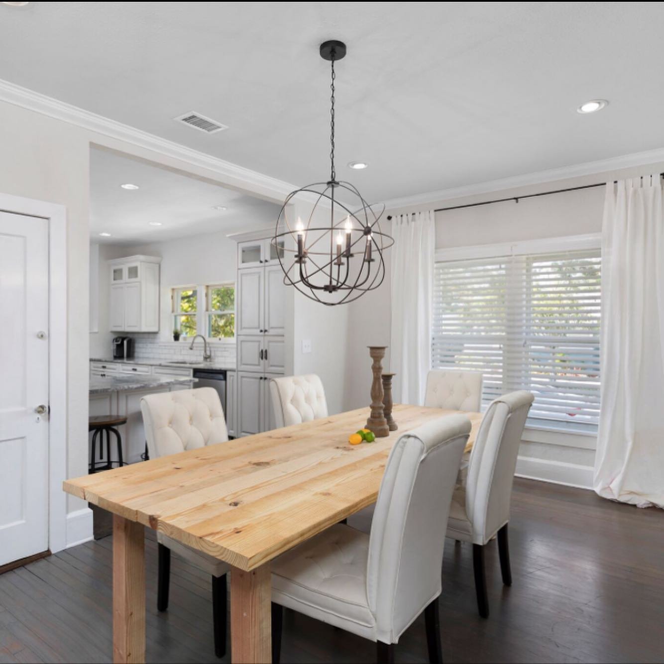 Dining room with a wooden table, white upholstered chairs, a modern chandelier, and a bright window with white curtains.