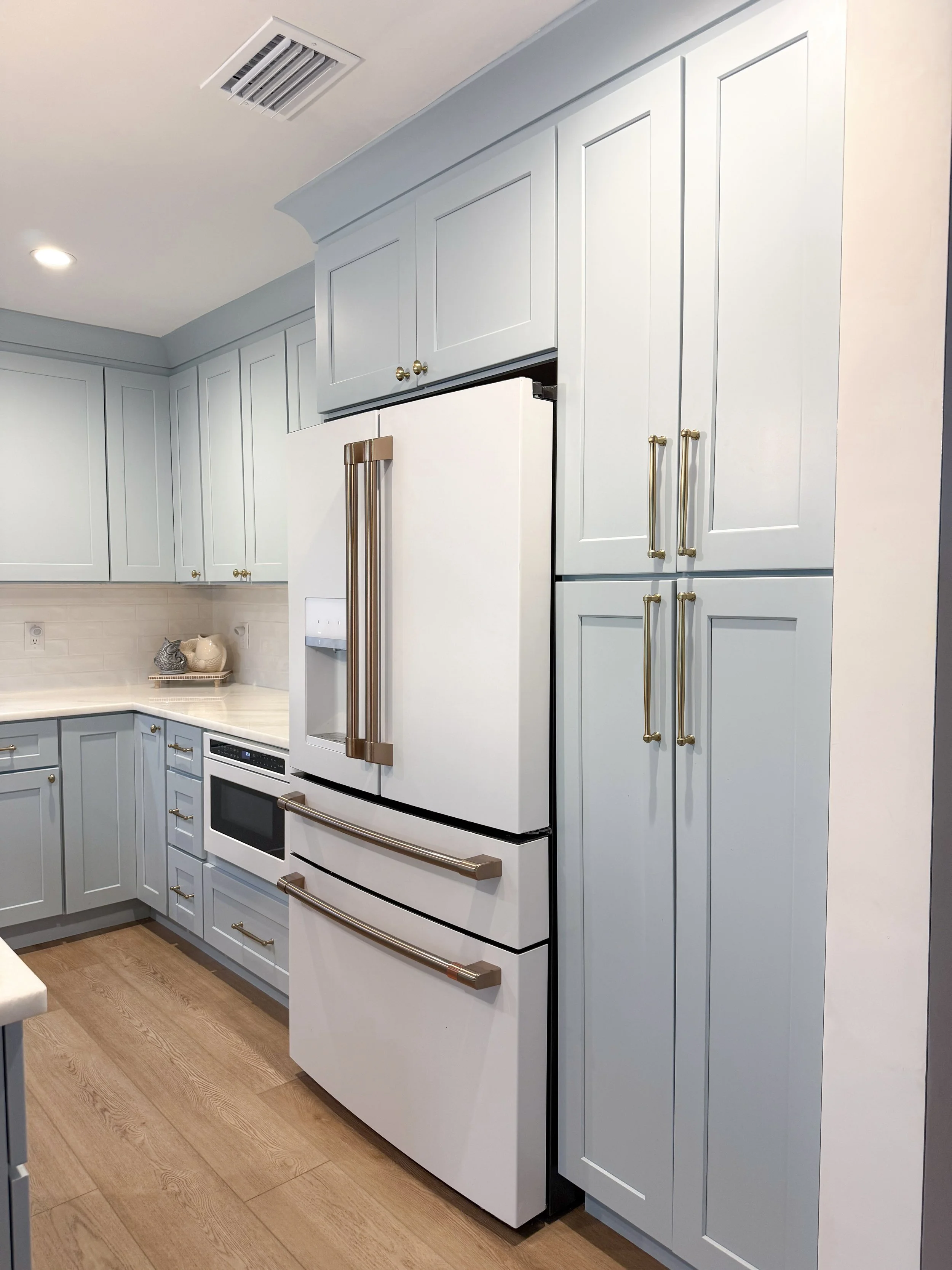 A modern kitchen with light blue cabinets, a white refrigerator with gold handles, a built-in microwave, and wooden flooring.