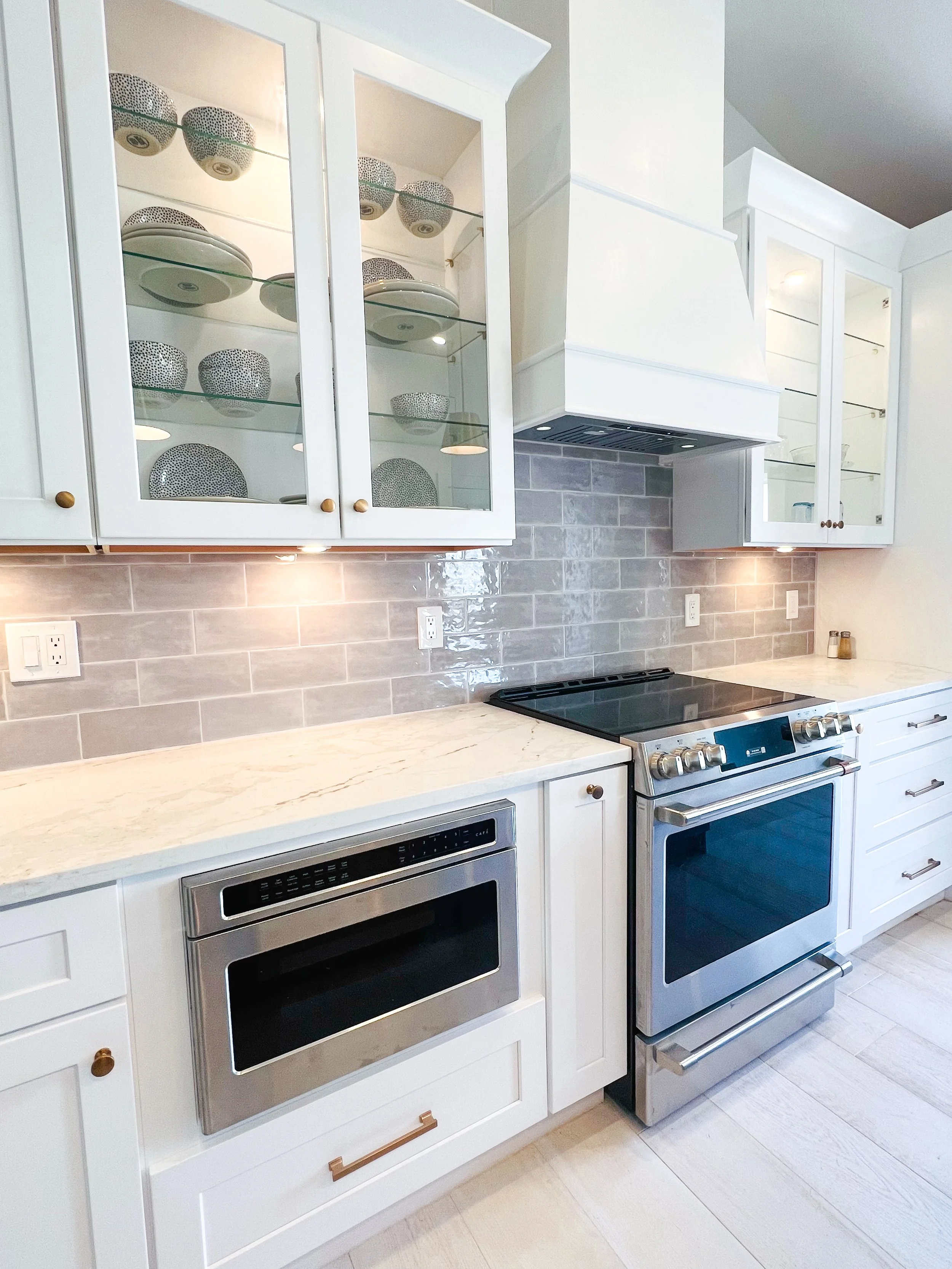 Modern kitchen with white cabinets, stainless steel oven and microwave, gray tile backsplash, marble countertop, glass-front cabinets with decorative bowls, and light wood flooring.
