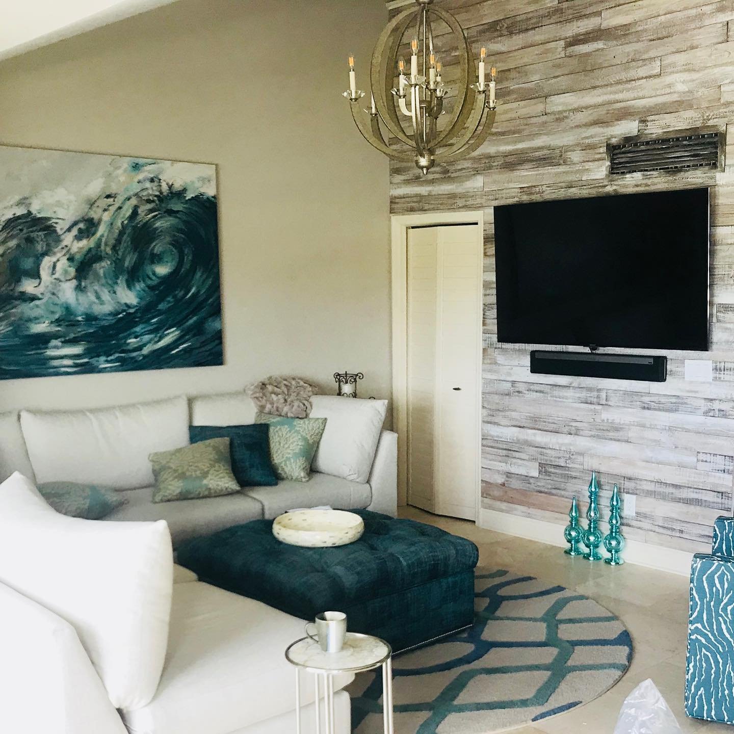 Living room with white sofa, teal and beige pillows, a black ottoman, wall-mounted TV on a wood plank wall, chandelier, abstract artwork, decorative teal vases, small side table with a mug, patterned rug.