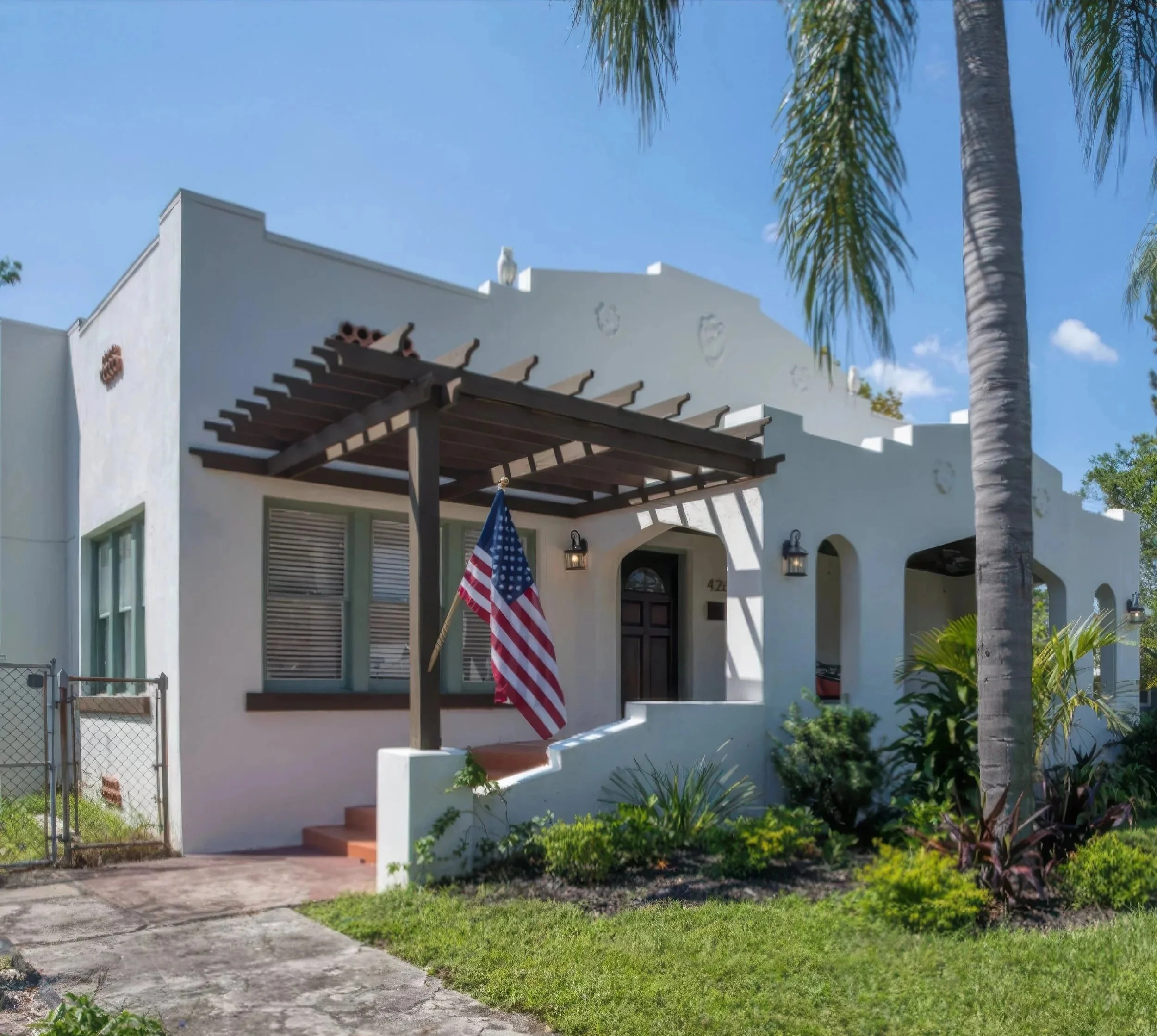 A white stucco house with a dark wooden pergola, American flag, and tropical landscaping under a blue sky.