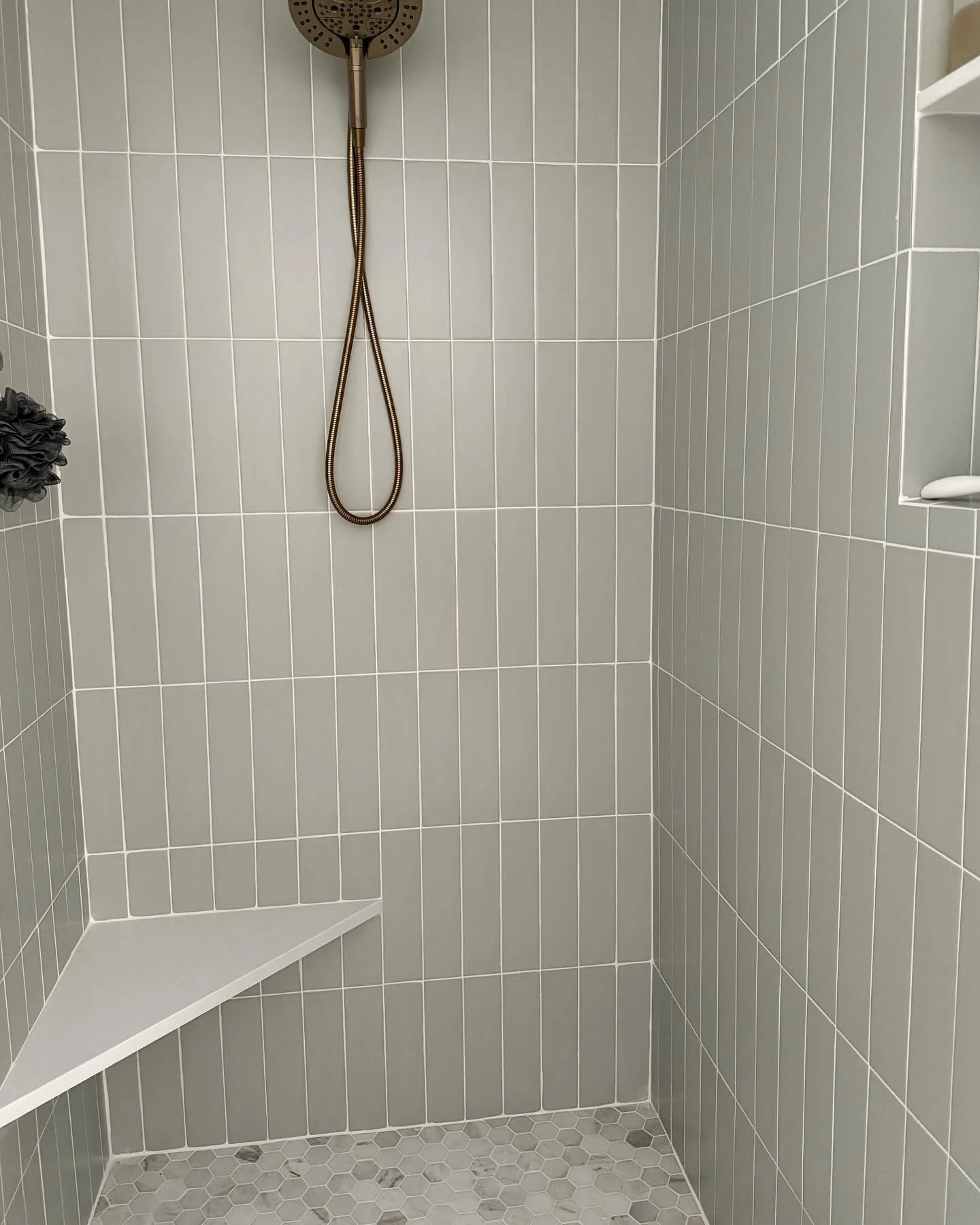 A modern shower with gray tiled walls, a round showerhead, a built-in corner shelf, and a hexagonal tile floor.