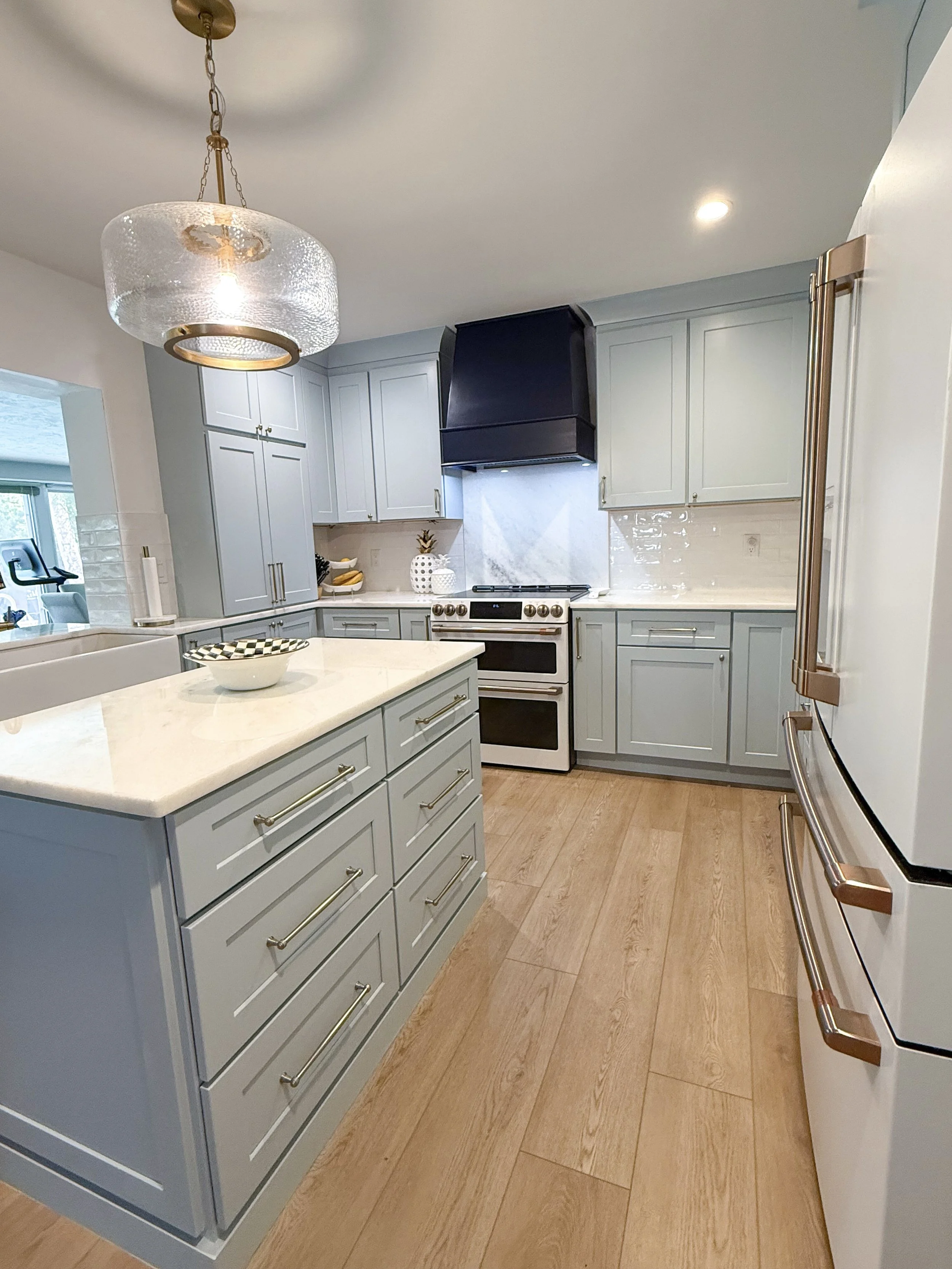 Modern kitchen with light blue cabinets, marble backsplash, stainless steel appliances, and a wooden floor. A hanging glass light fixture is above an island with a white countertop.