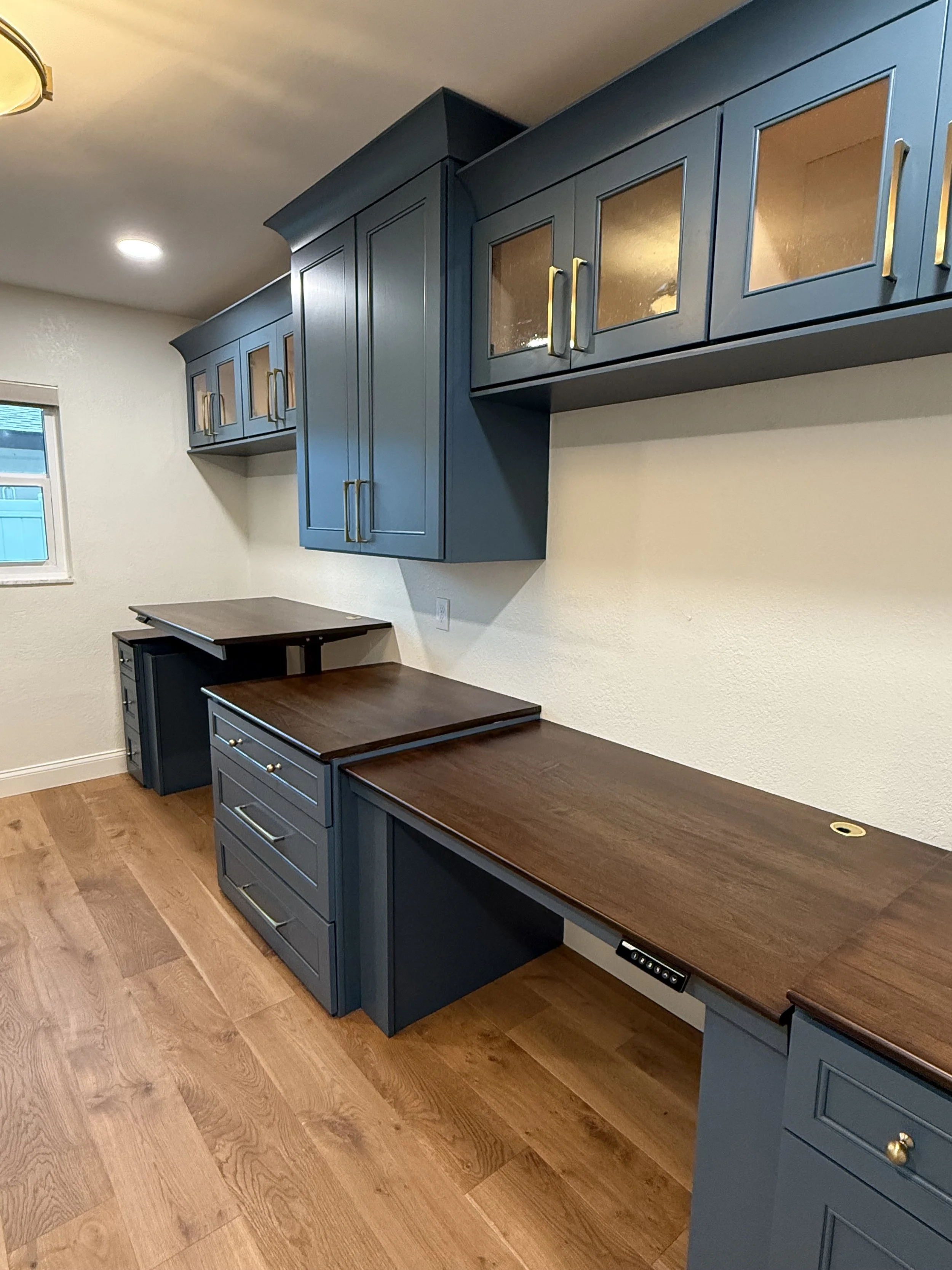 Custom blue cabinets with gold handles and brown wooden countertops in a home office or craft room with hardwood flooring.