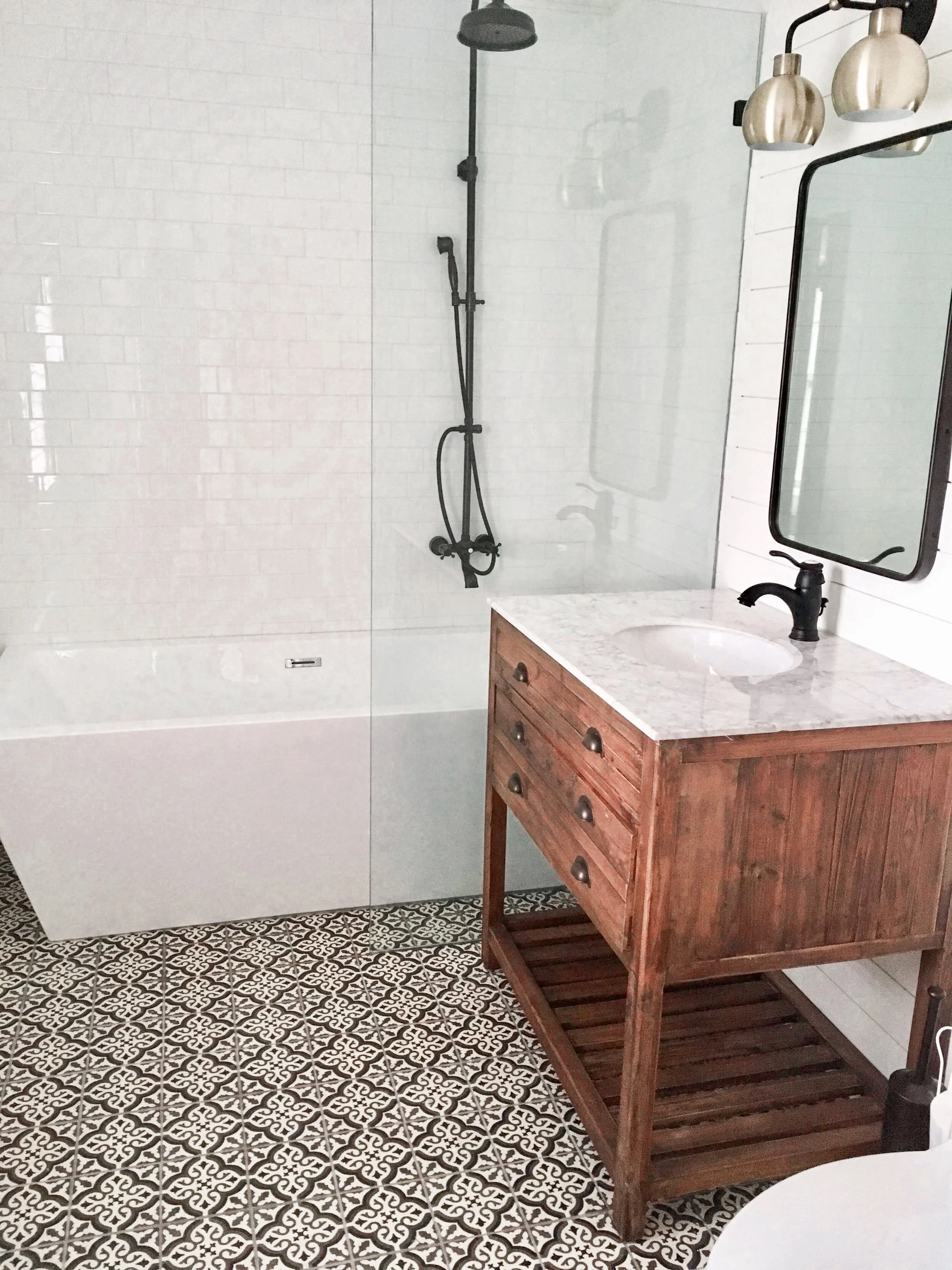 A bathroom featuring a shower with black fixtures, a white bathtub, a wooden vanity with a marble countertop and a black faucet, a mirror, and patterned floor tiles.
