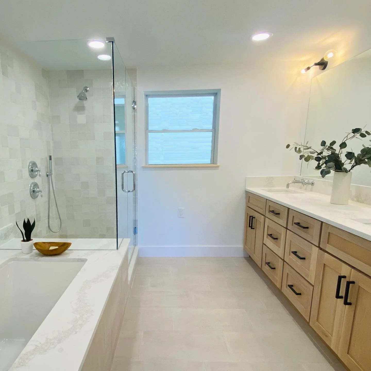Modern bathroom with a walk-in shower, light wood vanity with black handles, large mirror, potted plant, and a window