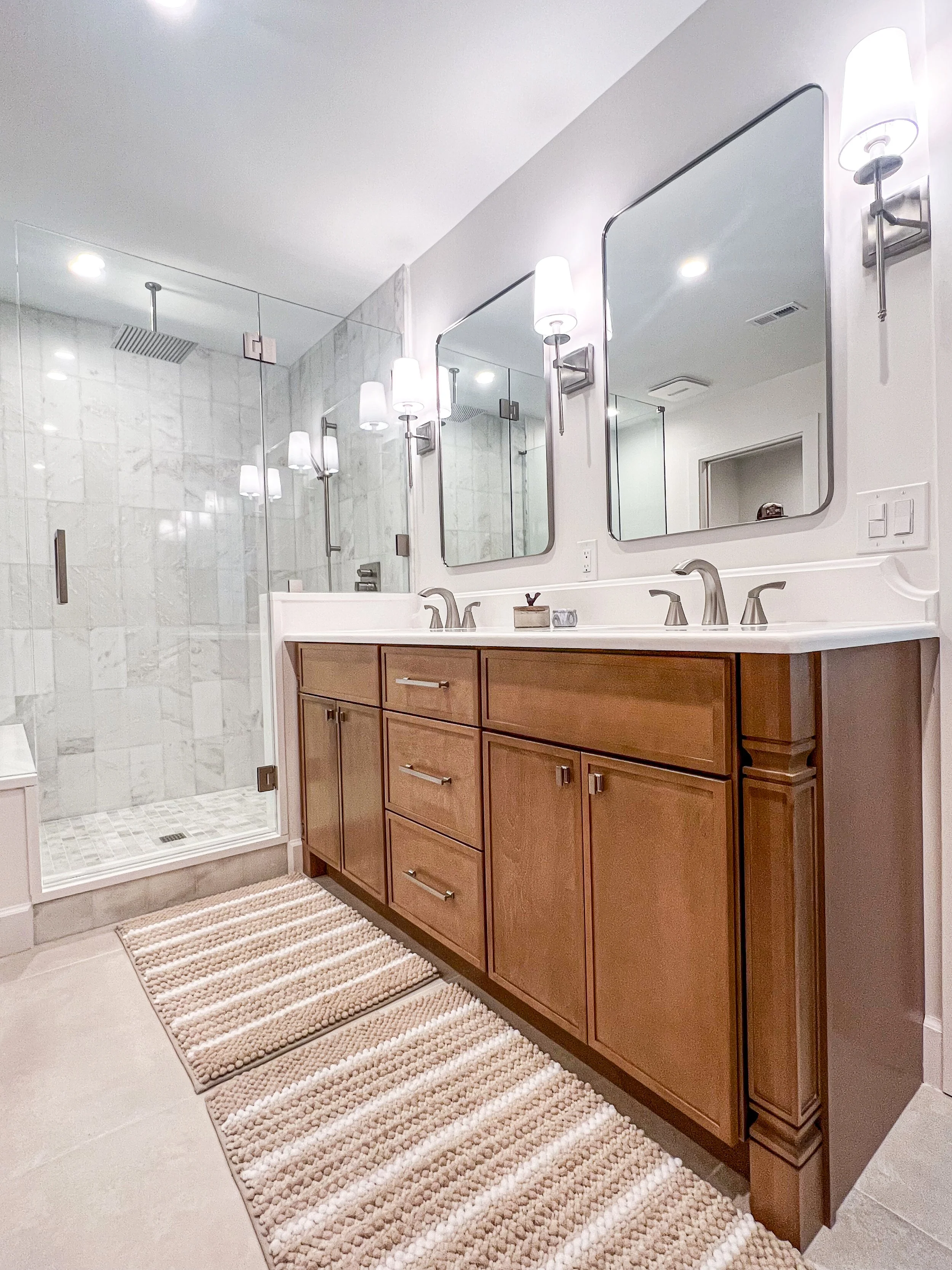 Modern bathroom with a dual-sink vanity, wooden cabinets, two large mirrors, wall sconces, a glass-enclosed shower with marble tiles and rainfall showerhead, and a beige striped rug on the tile floor.