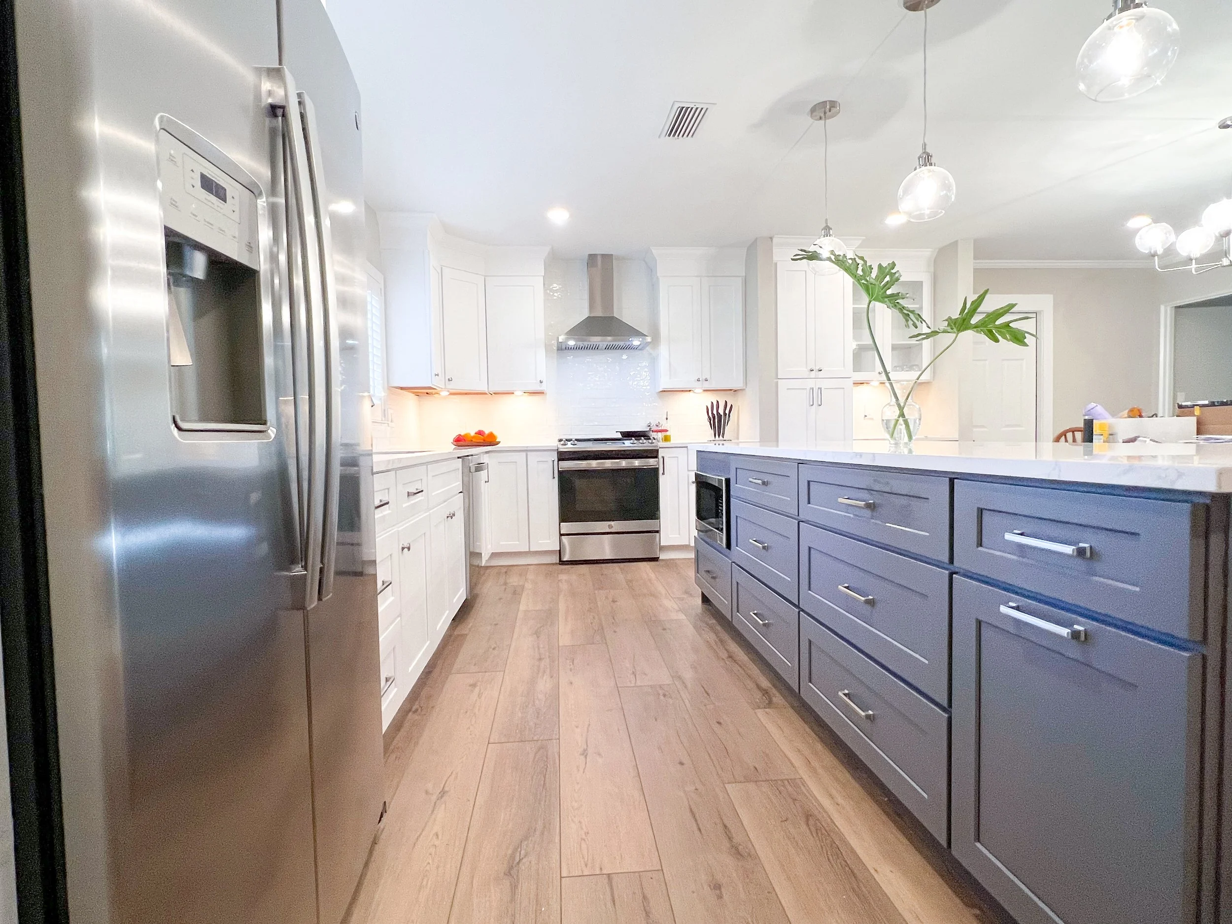 Modern kitchen with stainless steel refrigerator, white cabinets, and blue island with drawers, large vase with green leaves, and hardwood flooring.