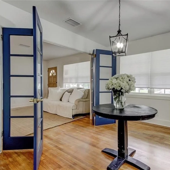 Interior view of a living room seen through blue folding doors, with a black round table holding a vase of white flowers, wood flooring, and a white sofa in the background.