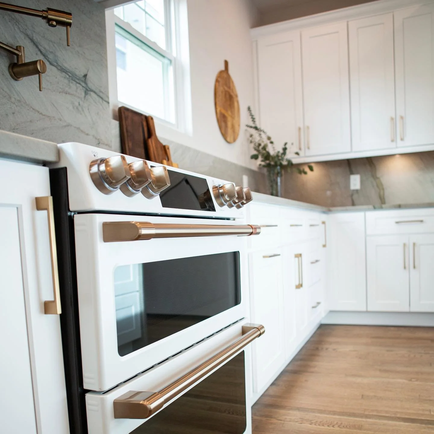 Modern kitchen with white cabinets, a marble countertop, and a white oven with stainless steel handles and knobs. There is a window above the countertop, hanging wooden cutting boards, and a vase with greenery on the counter. The flooring is wood.