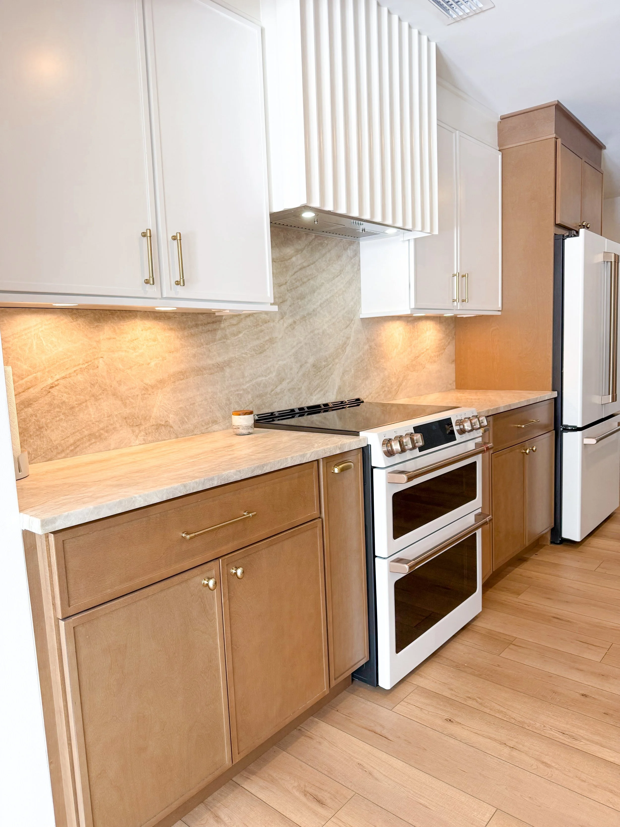 A kitchen with light wood lower cabinets, white upper cabinets, a beige marble backsplash, and a white oven with a black cooktop. Recessed lighting under the upper cabinets illuminates the countertop.