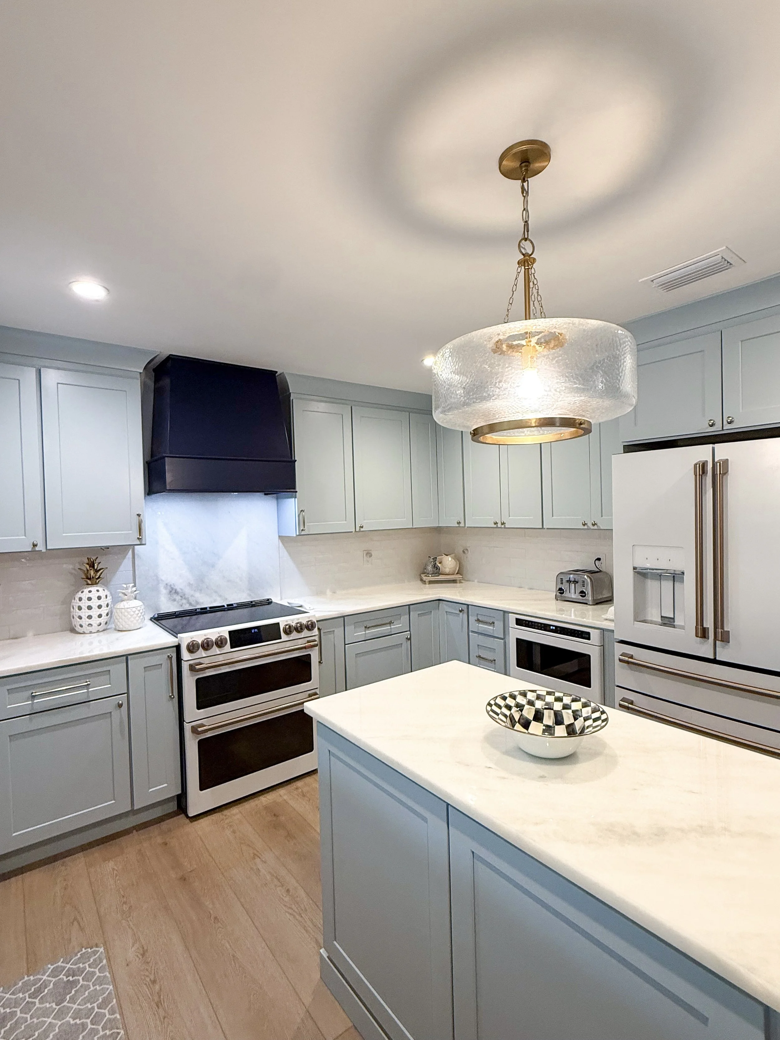 Modern kitchen with light blue cabinets, white marble countertops, and a central island with a checkered bowl. Features include a white stove, toaster, and a large refrigerator. A stylish glass and brass pendant light hangs from the ceiling.