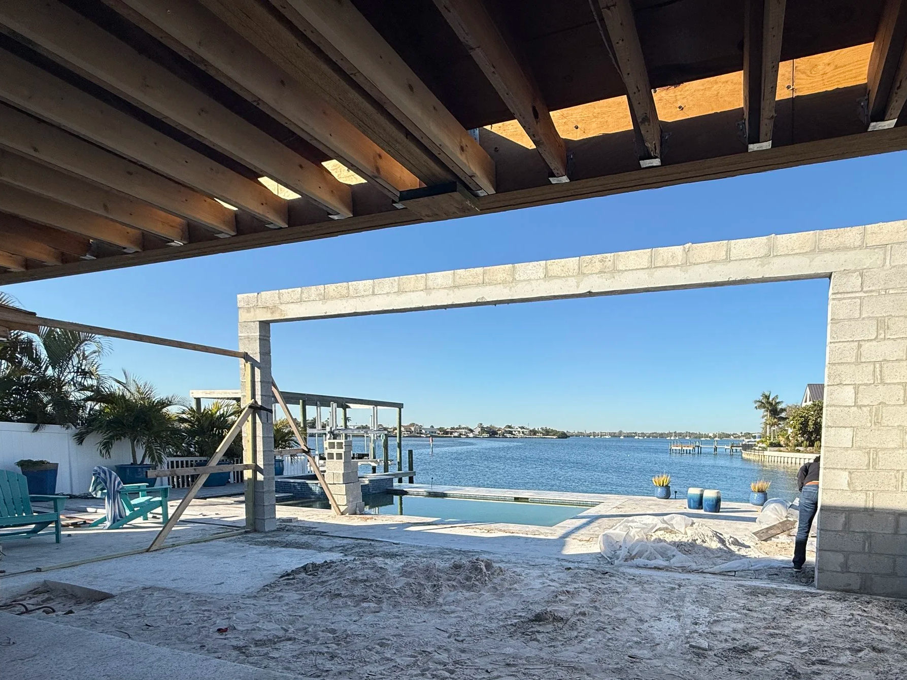 Construction site by water with partially built wall and outdoor furniture, overlooking a river or lake, under blue sky.