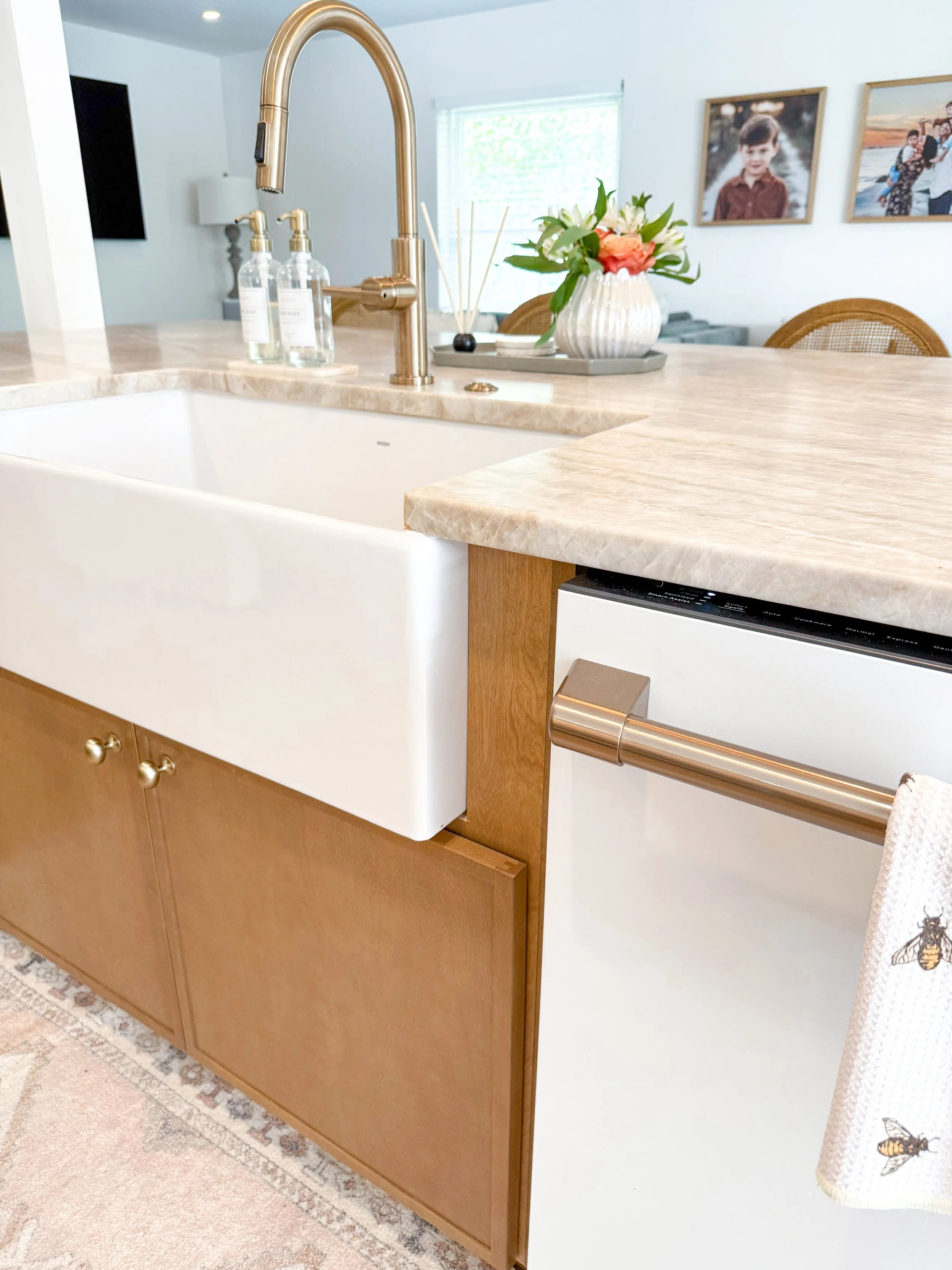 Close-up of a kitchen island with a white farmhouse sink, a gold faucet, a beige marble countertop, a flower arrangement, and a dishwasher with a towel hanging on its handle.