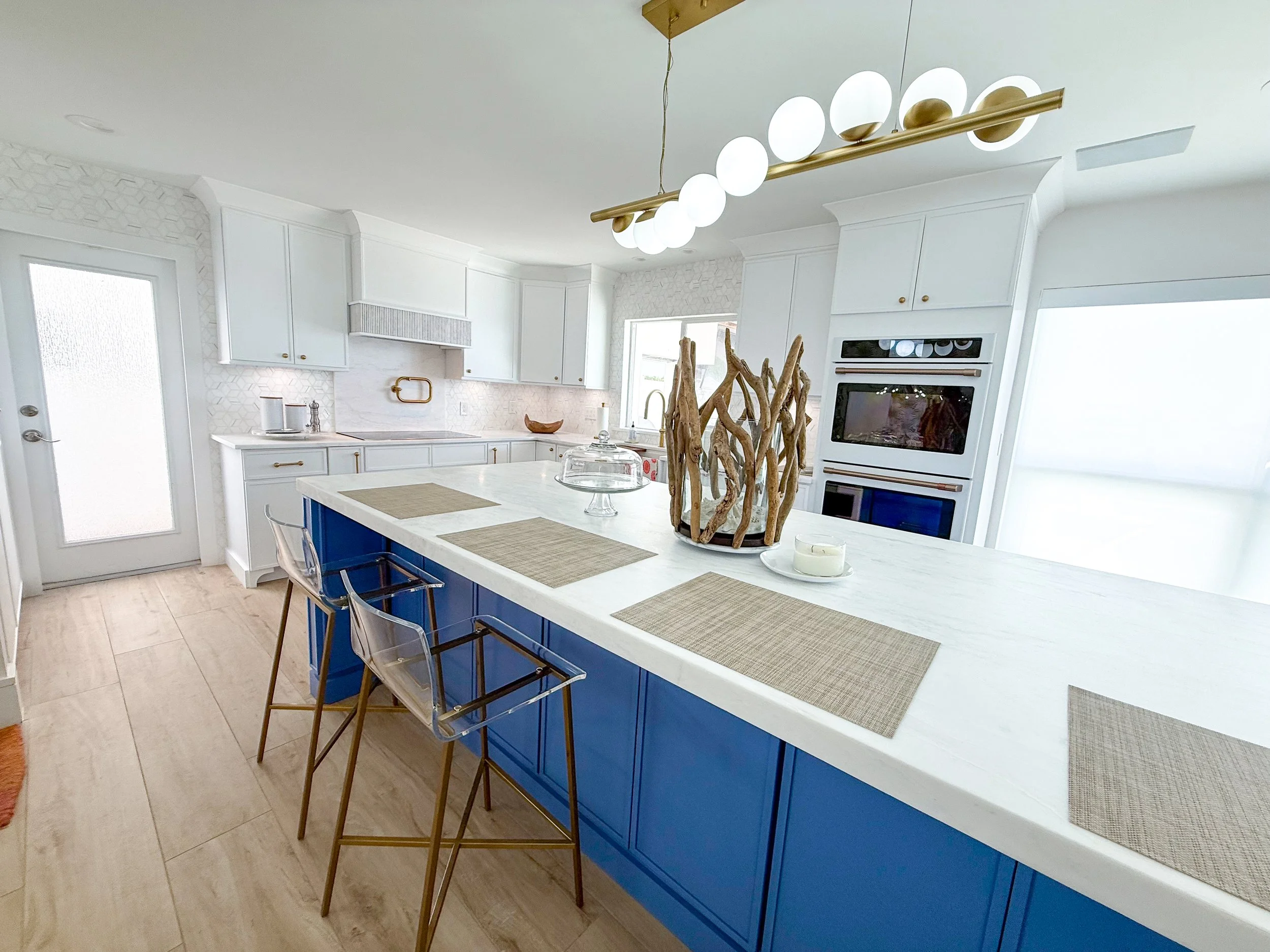 Modern kitchen with white cabinets, a large white island with blue lower cabinets, two transparent bar stools with gold legs, placed on a beige wooden floor. The kitchen features built-in ovens, a window with white curtains, and a contemporary black 