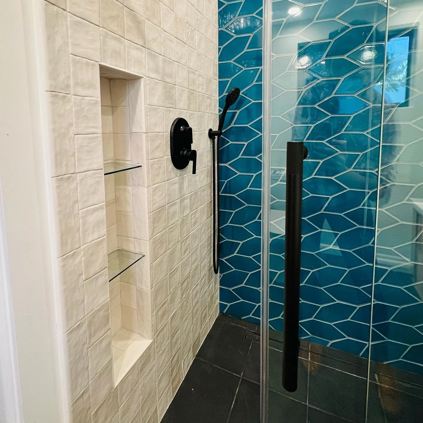 Shower with beige tiled wall, built-in shelf, and a glass door with black handle; blue hexagonal tiled wall behind the shower with a black handheld showerhead.