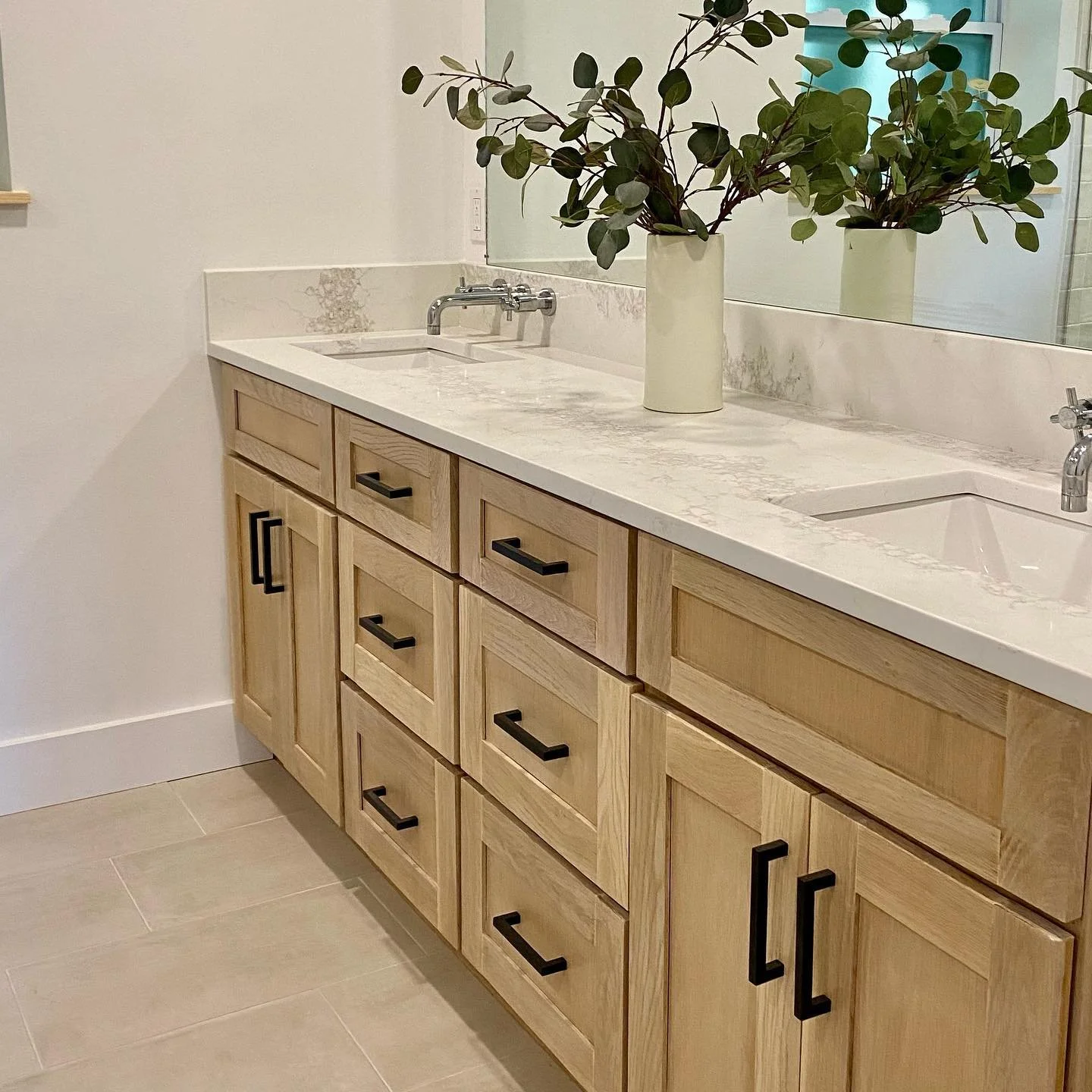 Bathroom vanity with light wood cabinets, black handles, a marble countertop, two sinks, a large mirror, and two potted plants.