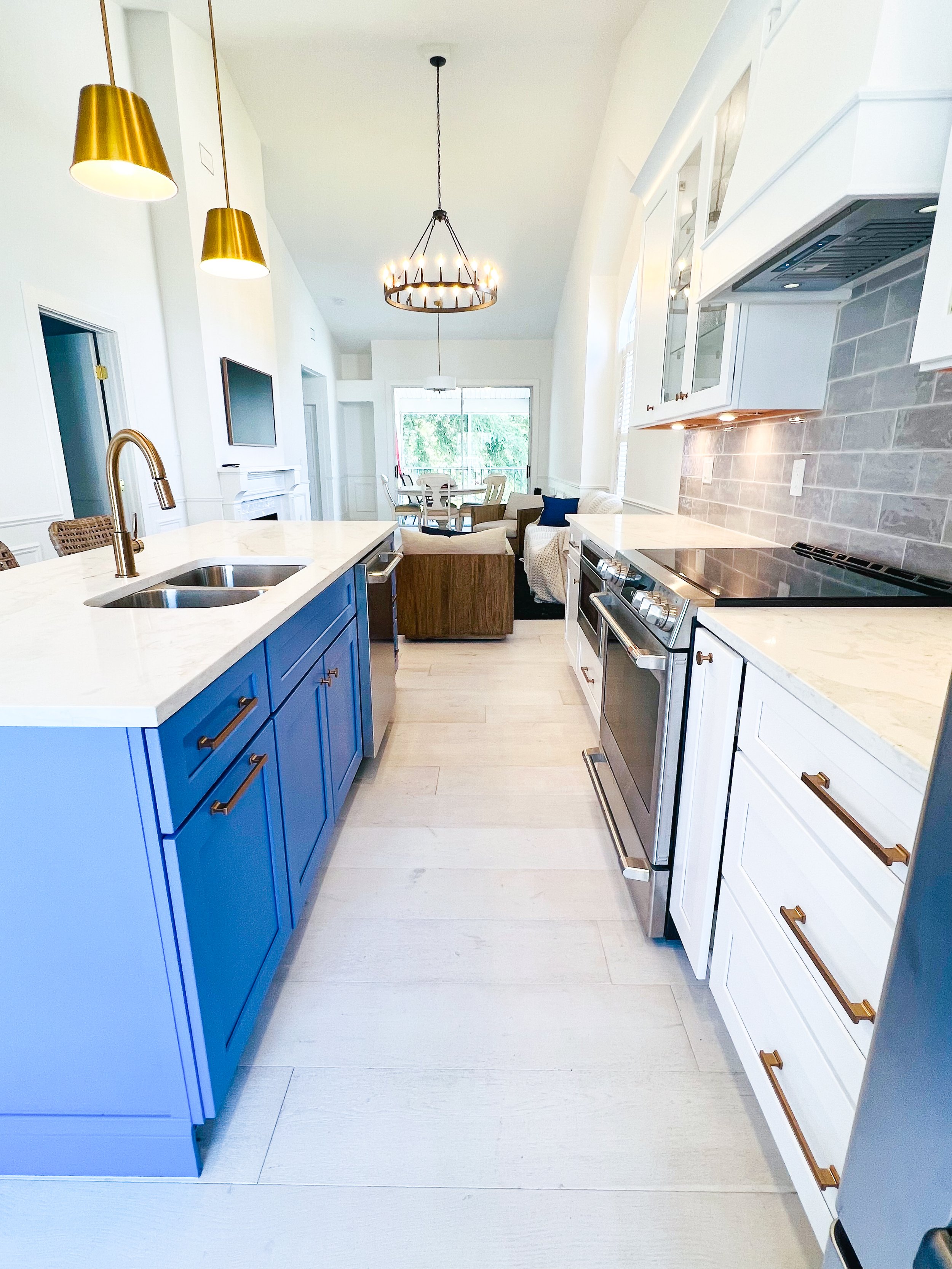 Modern kitchen with blue and white cabinets, stainless steel appliances, gold cabinet handles, pendant lights, and a view of a dining and living area with large windows.