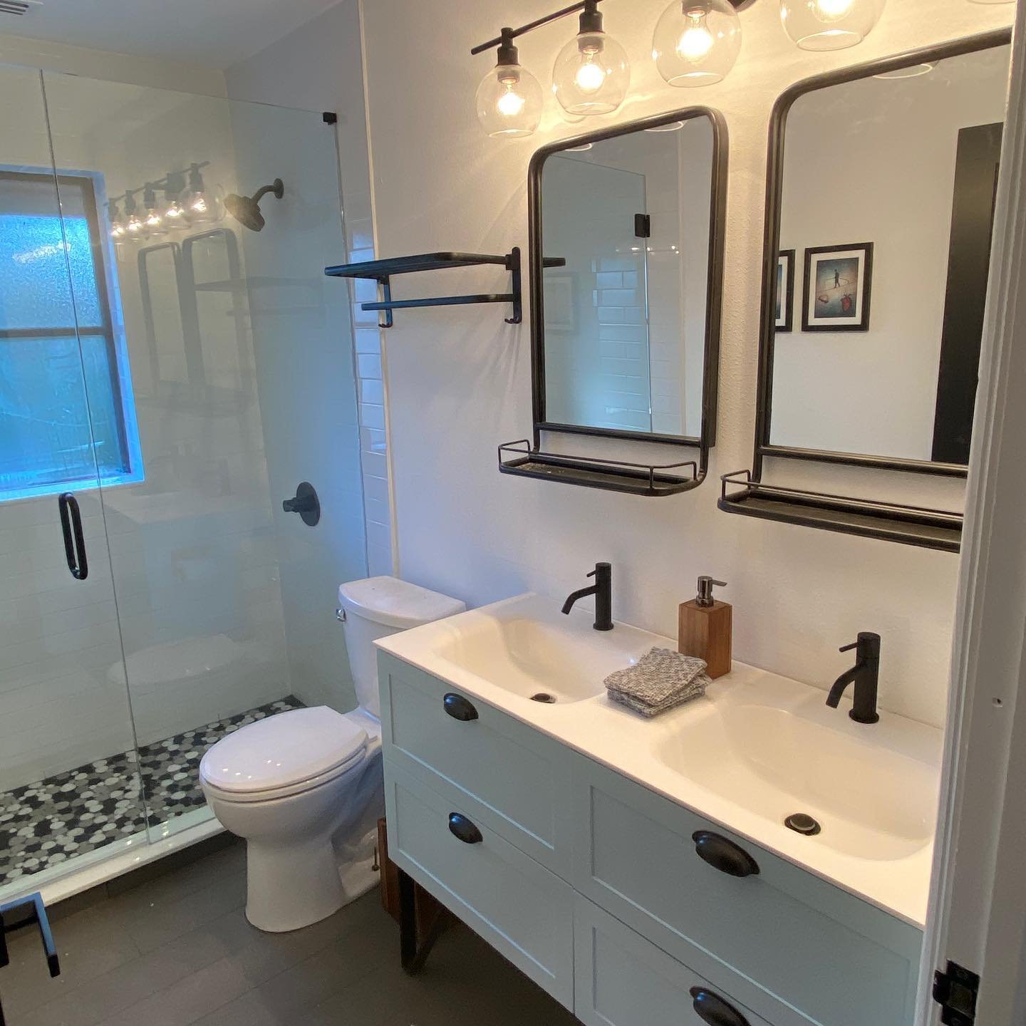 Modern bathroom with a double sink vanity, black fixtures, and a shower with a glass door and pebble tile floor. Two mirrors are mounted above the sinks, and above them are three hanging lightbulbs with glass shades. There is a small window in the sh