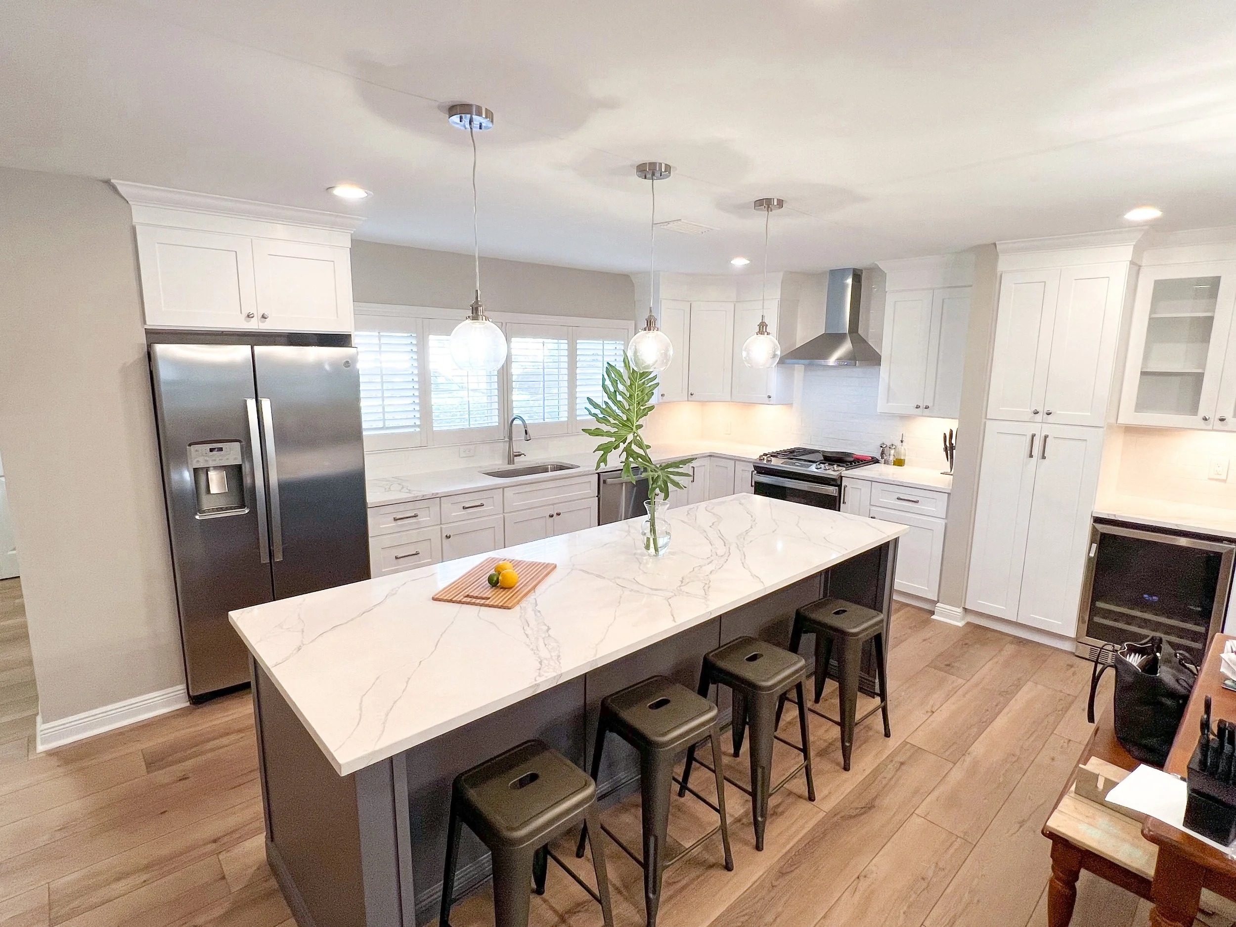 Modern kitchen with white cabinetry, marble countertop island, stainless steel refrigerator, and hanging pendant lights. Wooden floor with bar stools at the island and a window with shutters above the sink.