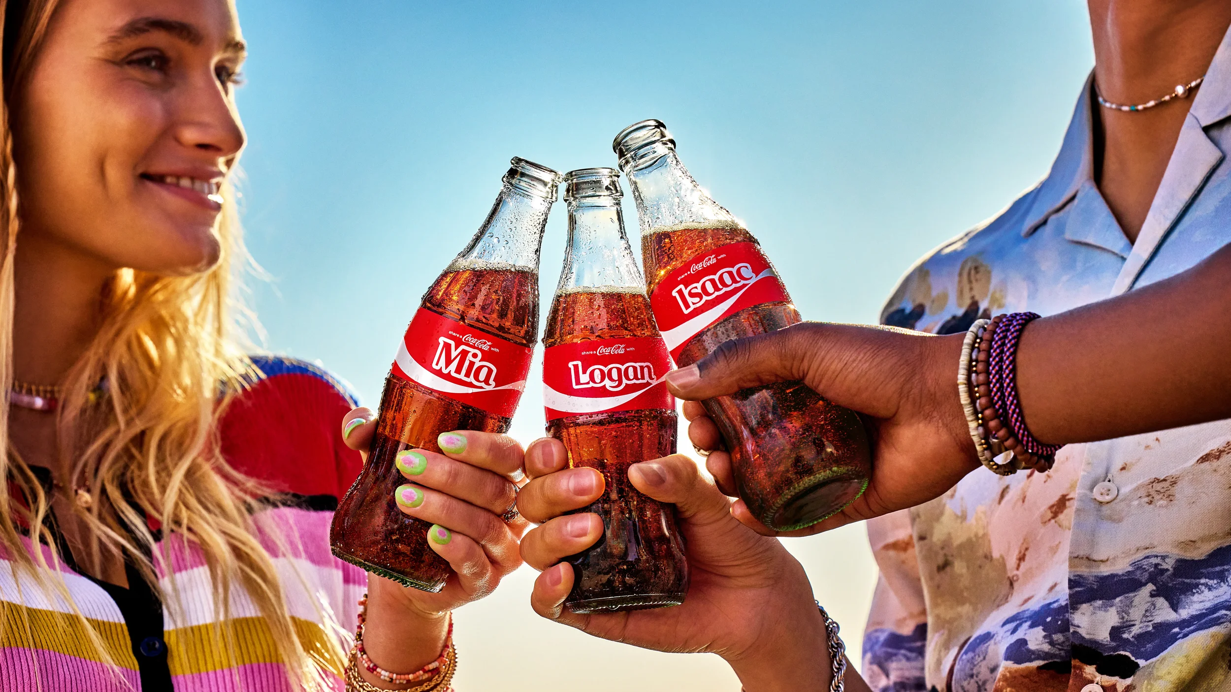  Three glass Coca-Cola bottles with personalized labels being clinked together outdoors under bright sunlight. 