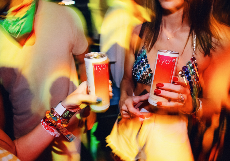  Two people holding cans of Iyo functional drink at a vibrant party scene with colorful lights and festival wristbands, highlighting a lively social atmosphere. 