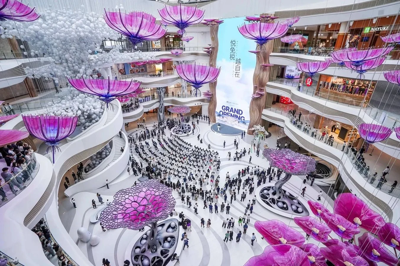  Grand opening event in a modern airport atrium featuring large pink floral installations, hanging decorations, and crowds gathered around a central stage with a tall digital screen. 