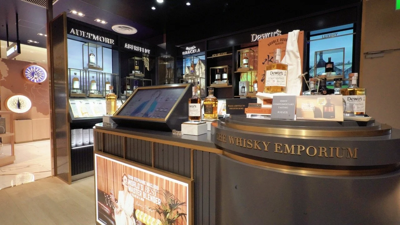  The Whisky Emporium airport retail display featuring premium Scotch brands like Dewar’s and Aultmore, interactive tasting station, and elegant dark wood shelving with illuminated bottle showcases. 