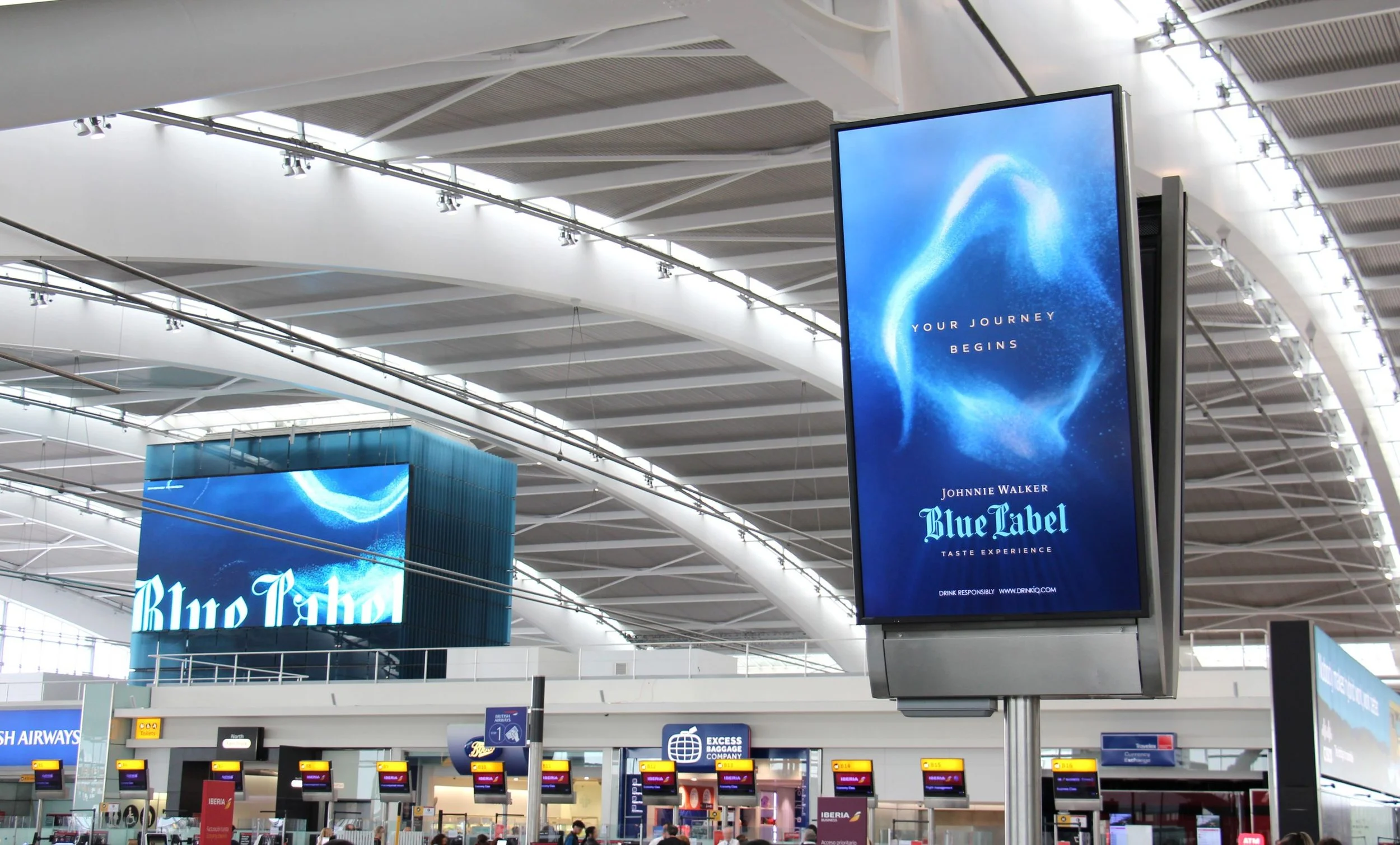  Johnnie Walker Blue Label digital advertisement displayed in a modern airport terminal with curved architectural ceiling, large screens, and check-in counters in the background. 