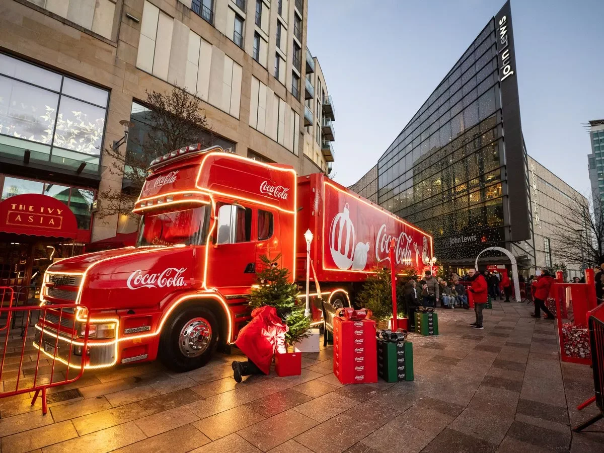 conic Coca-Cola Christmas truck illuminated with festive lights outside John Lewis, surrounded by holiday decorations, gift boxes, and visitors enjoying the seasonal celebration in a city shopping district. 