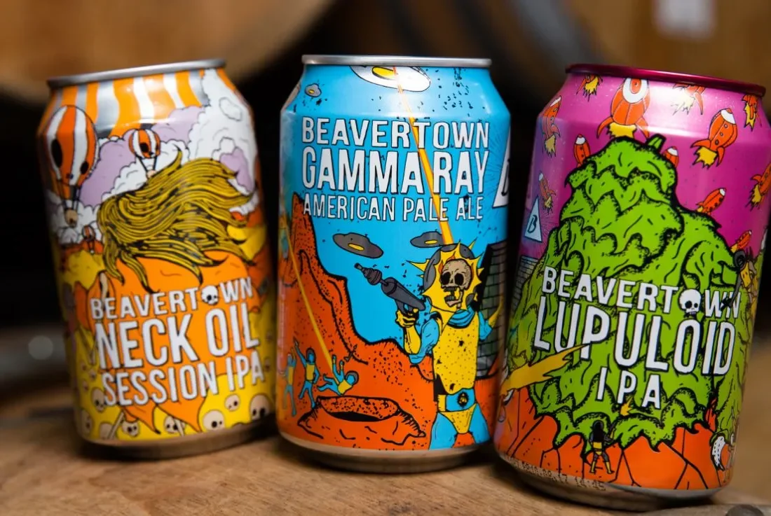  Three colorful Beavertown beer cans displayed on a wooden surface, featuring Neck Oil Session IPA, Gamma Ray American Pale Ale, and Lupuloid IPA with vibrant, illustrated designs. 