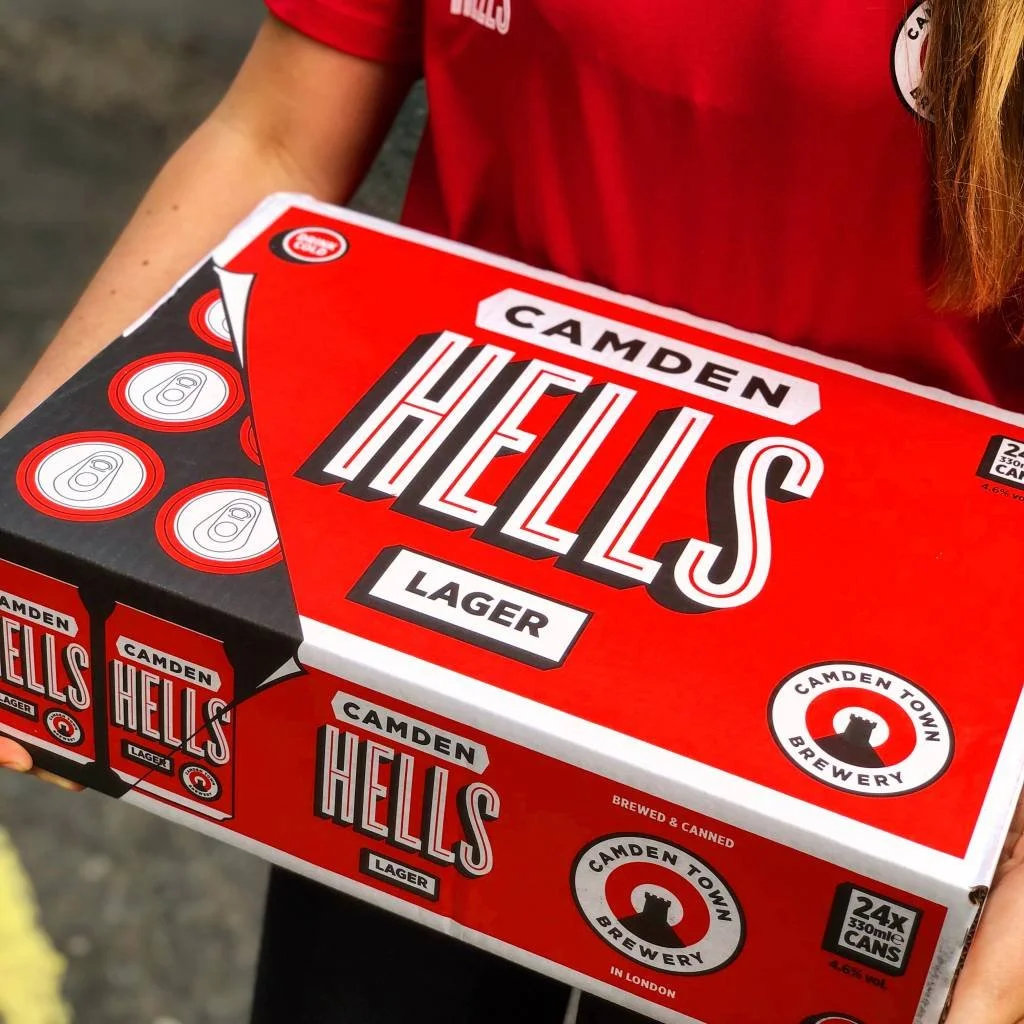  Person holding a bright red Camden Hells Lager 24-pack box from Camden Town Brewery, featuring bold white and black branding and logo design. 