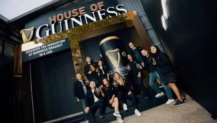  Group of people posing in front of the House of Guinness entrance, featuring a large Guinness Draught can display and branded signage promoting Guinness innovation. 