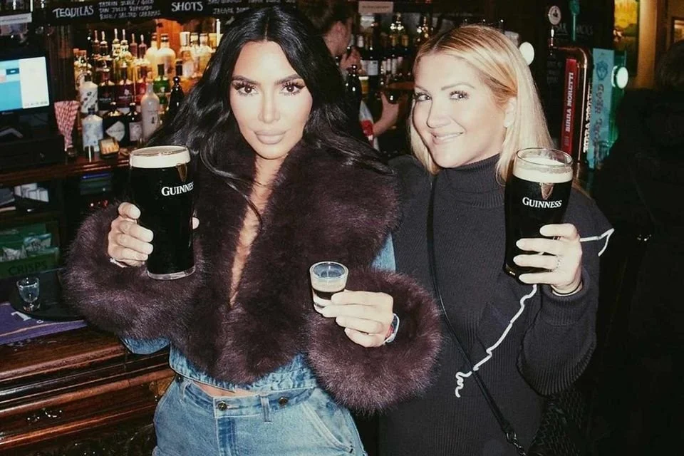  Kim Kardashian at a bar holding pints of Guinness stout and a shot glass, with shelves of liquor bottles and drink menus visible in the background. 
