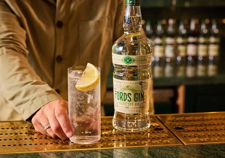  Fords Gin bottle on a bar beside a gin and tonic with lemon, captured in a warm cocktail bar setting. 