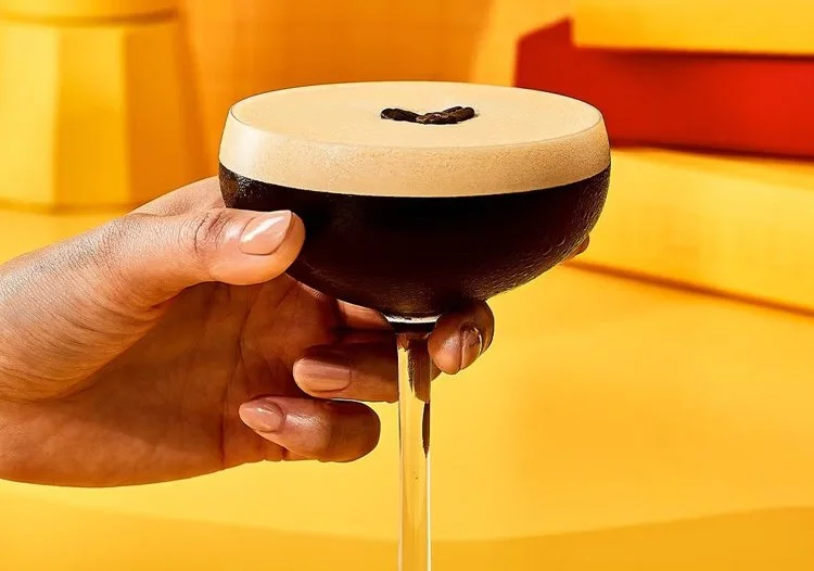  Hand holding a fresh espresso martini garnished with coffee beans against a bold yellow background. 