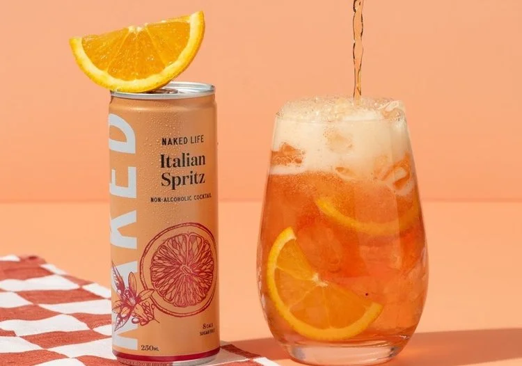  Naked Life Italian Spritz can next to a glass filled with orange slices and a poured non‑alcoholic spritz on a bright summer backdrop. 
