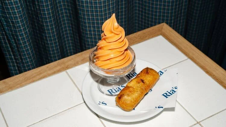  Ria’s Ice Cream x Big Kid Ice Cream dessert featuring orange soft‑serve in a glass dish paired with a golden fried pastry on branded Ria’s paper, displayed on a tiled countertop. 