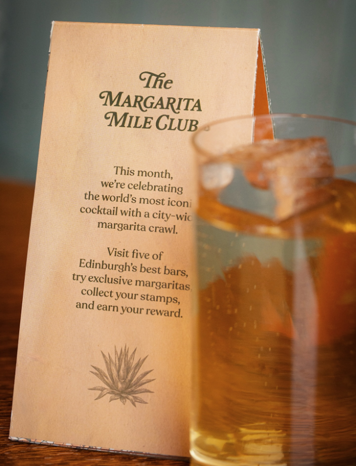  Tabletop display promoting The Margarita Mile Club in Edinburgh, featuring a printed event card with agave illustration beside a classic margarita cocktail served over ice on a wooden surface. 