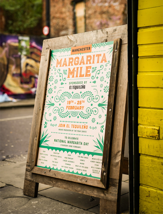  Street‑side signboard promoting Margarita Mile in Manchester, sponsored by El Tequileño, featuring illustrated agave motifs and event details celebrating National Margarita Day. 