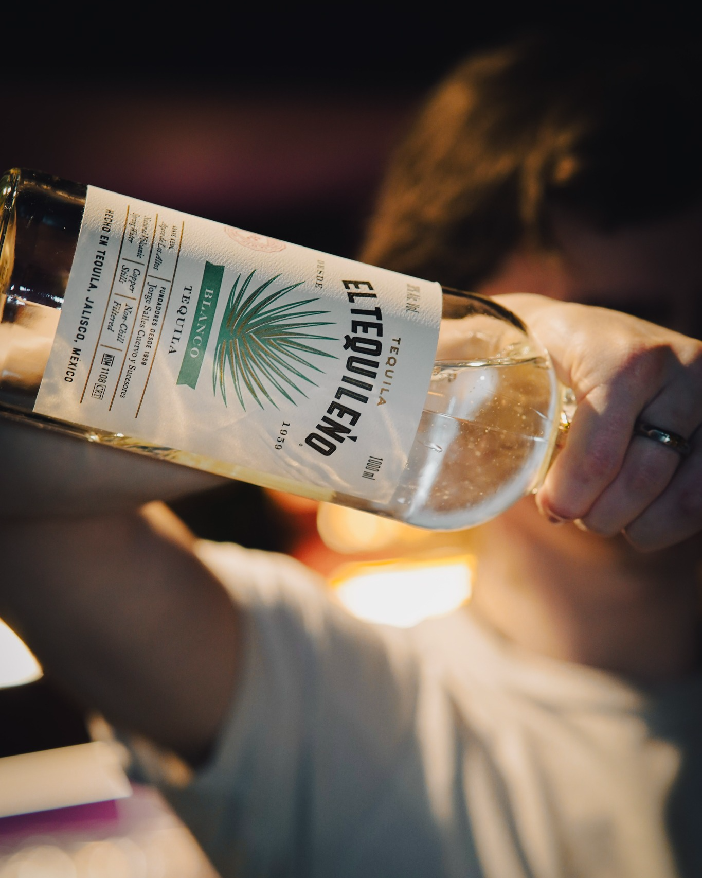  El Tequileño tequila bottle being poured behind a bar, showcasing the agave‑inspired label and premium clear spirit, captured in warm, atmospheric lighting. 
