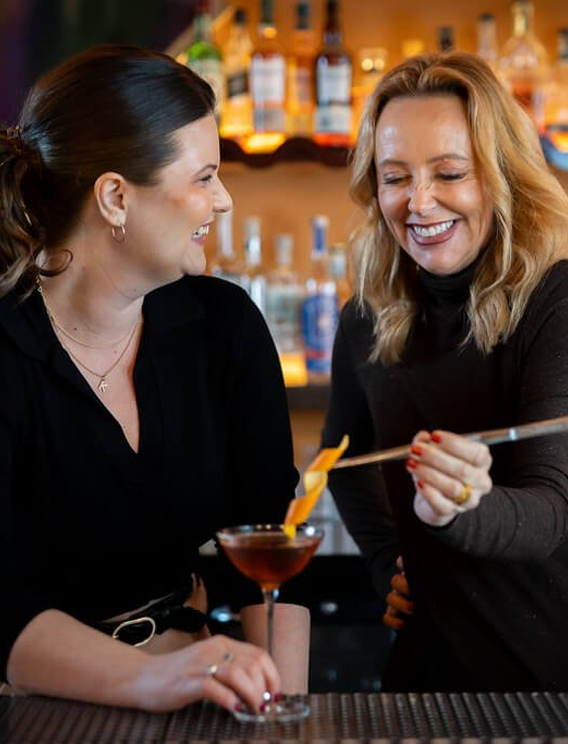  Two bartenders preparing a cocktail behind a bar, garnishing a coupe glass with citrus peel, with illuminated shelves of spirits in the background creating a warm, premium bar setting. 