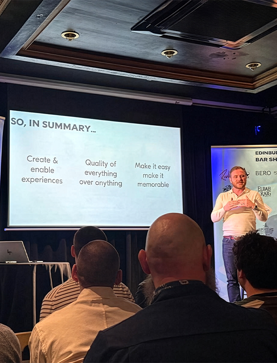  Speaker presenting a summary slide at the Edinburgh Bar Show, with key points on creating experiences, quality and memorability displayed on a large screen to a seated drinks industry audience. 