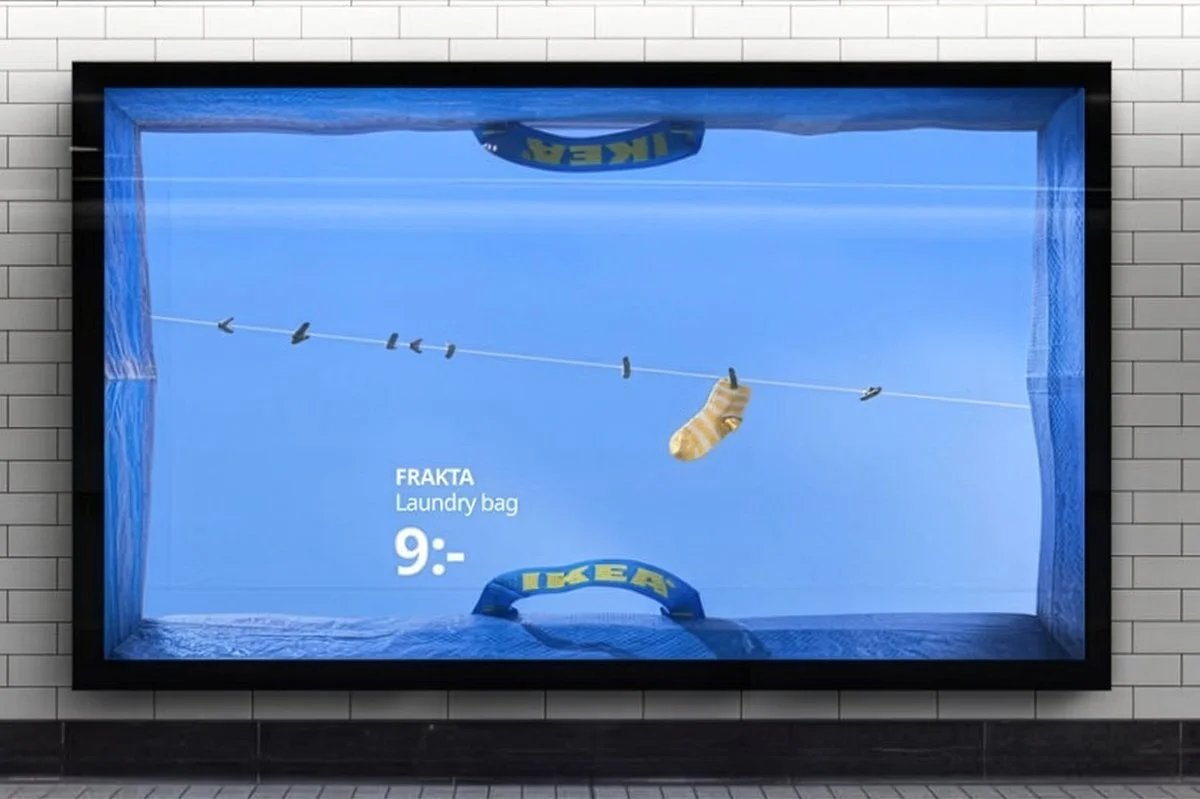  IKEA FRAKTA campaign billboard featuring a blue bag framed as a laundry scene with a clothesline and sock, promoting the FRAKTA laundry bag priced at 9, displayed on a tiled wall. 